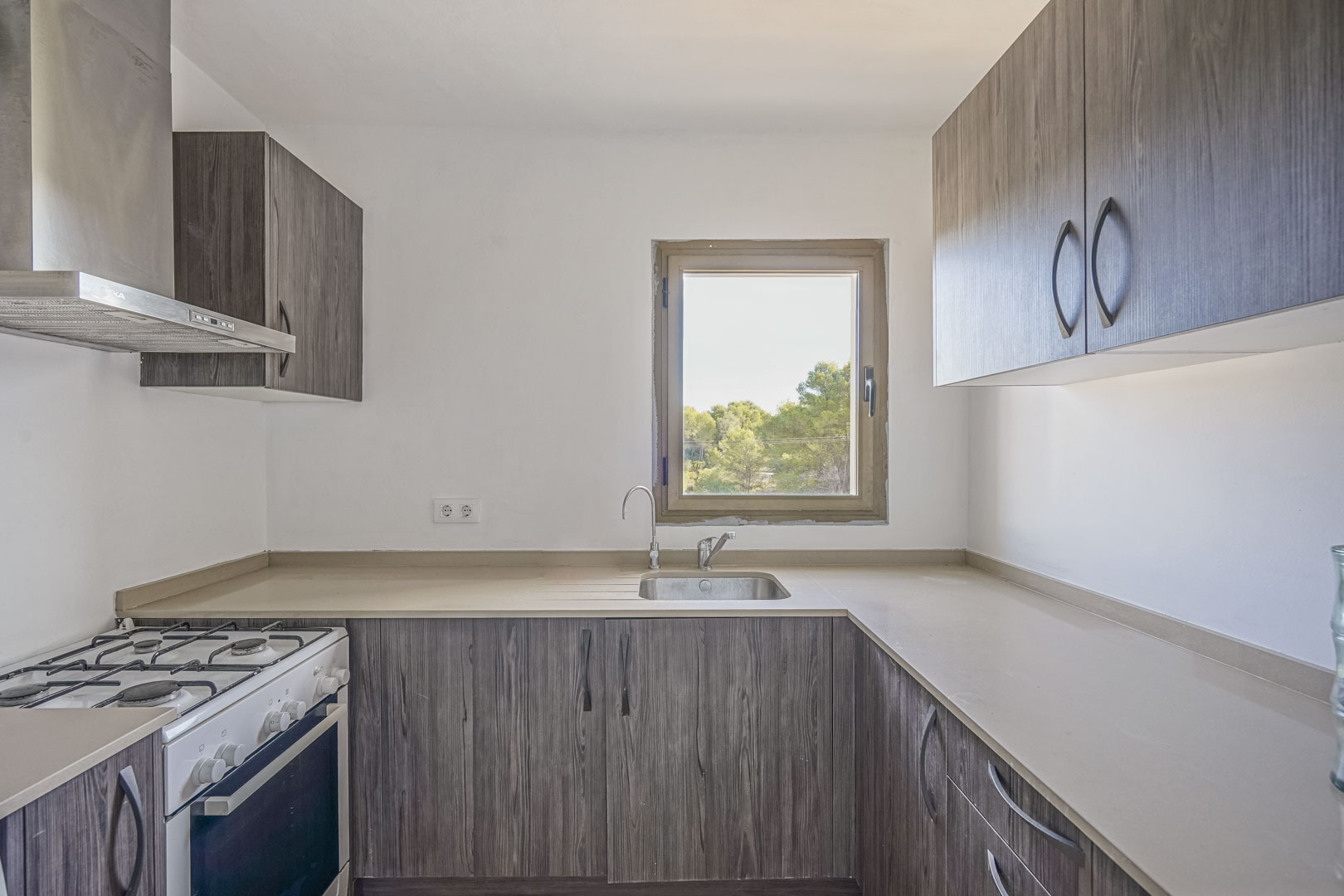 Cocina funcional con armarios de efecto madera oscura, encimera de tono neutro y ventana con vistas al exterior. Incluye cocina de gas y campana extractora de acero inoxidable.