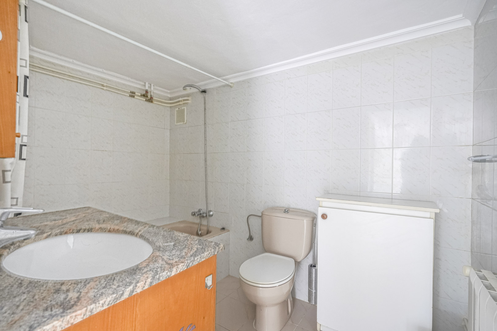 Cuarto de baño con encimera de granito, lavabo encastrado, inodoro en tono beige y paredes revestidas con azulejos blancos. Incluye zona de ducha y radiador lateral.