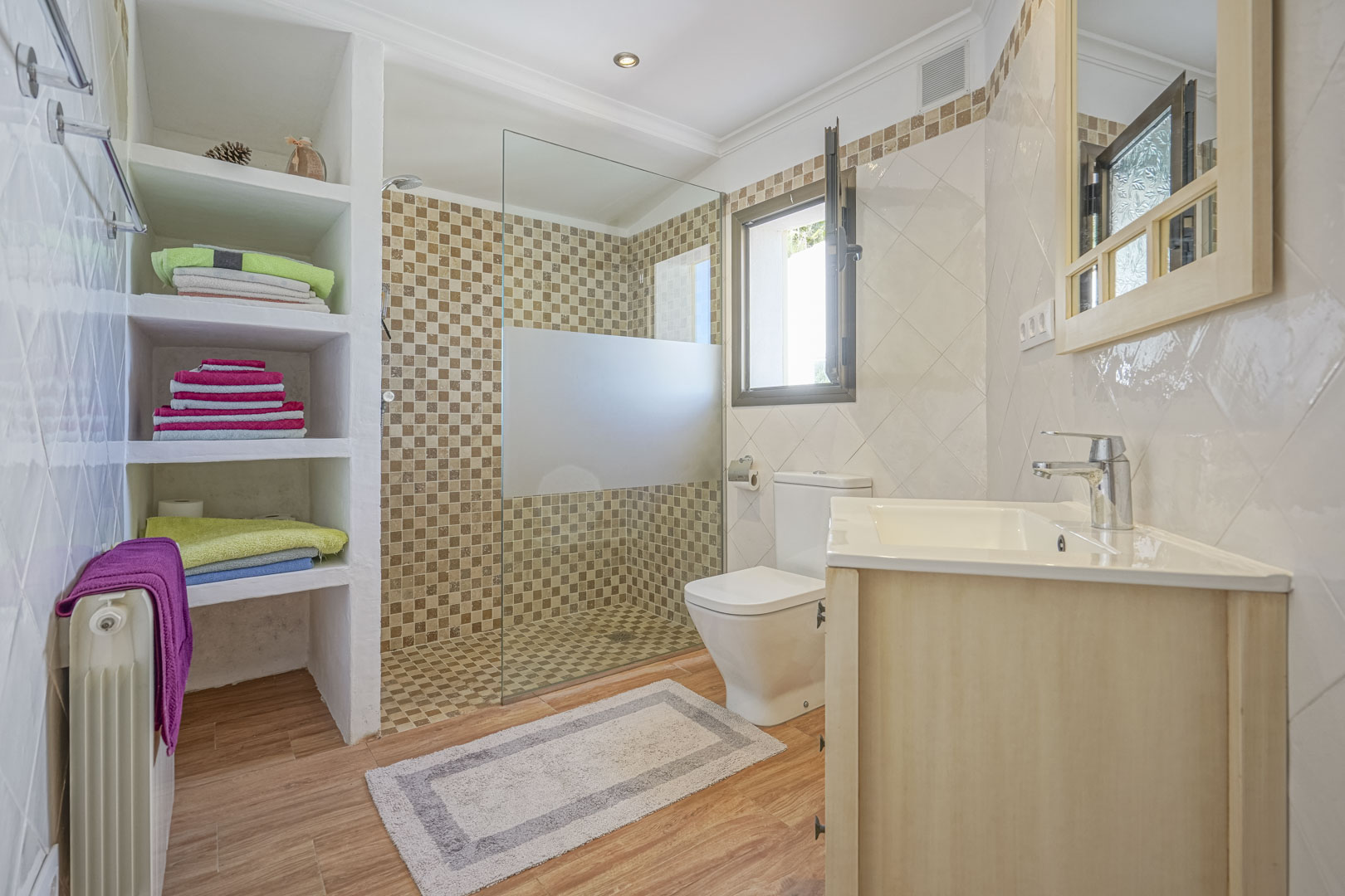 Cuarto de baño renovado con ducha de obra en mosaico, mampara de cristal, estantería integrada de obra, suelo de gres efecto madera y mueble de lavabo en madera clara.