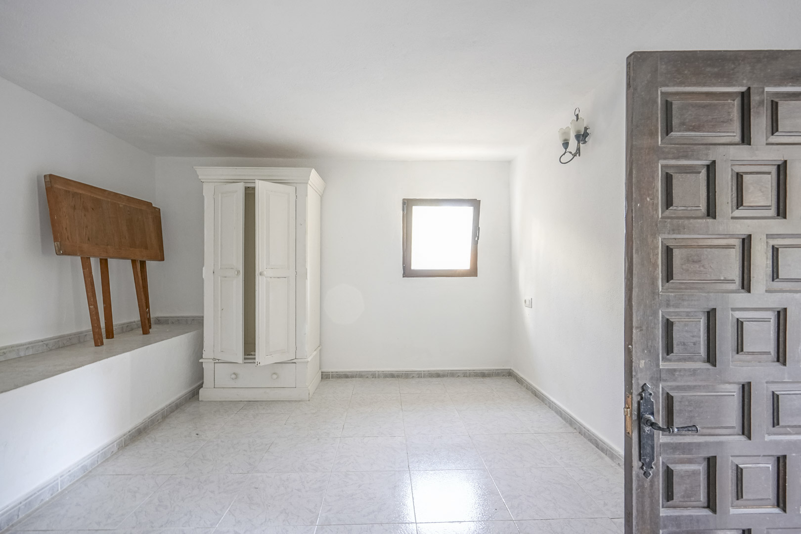 Habitación con suelos cerámicos claros, paredes blancas y luz natural. Incluye una puerta de madera tallada tradicional y un armario exento de estilo clásico.
