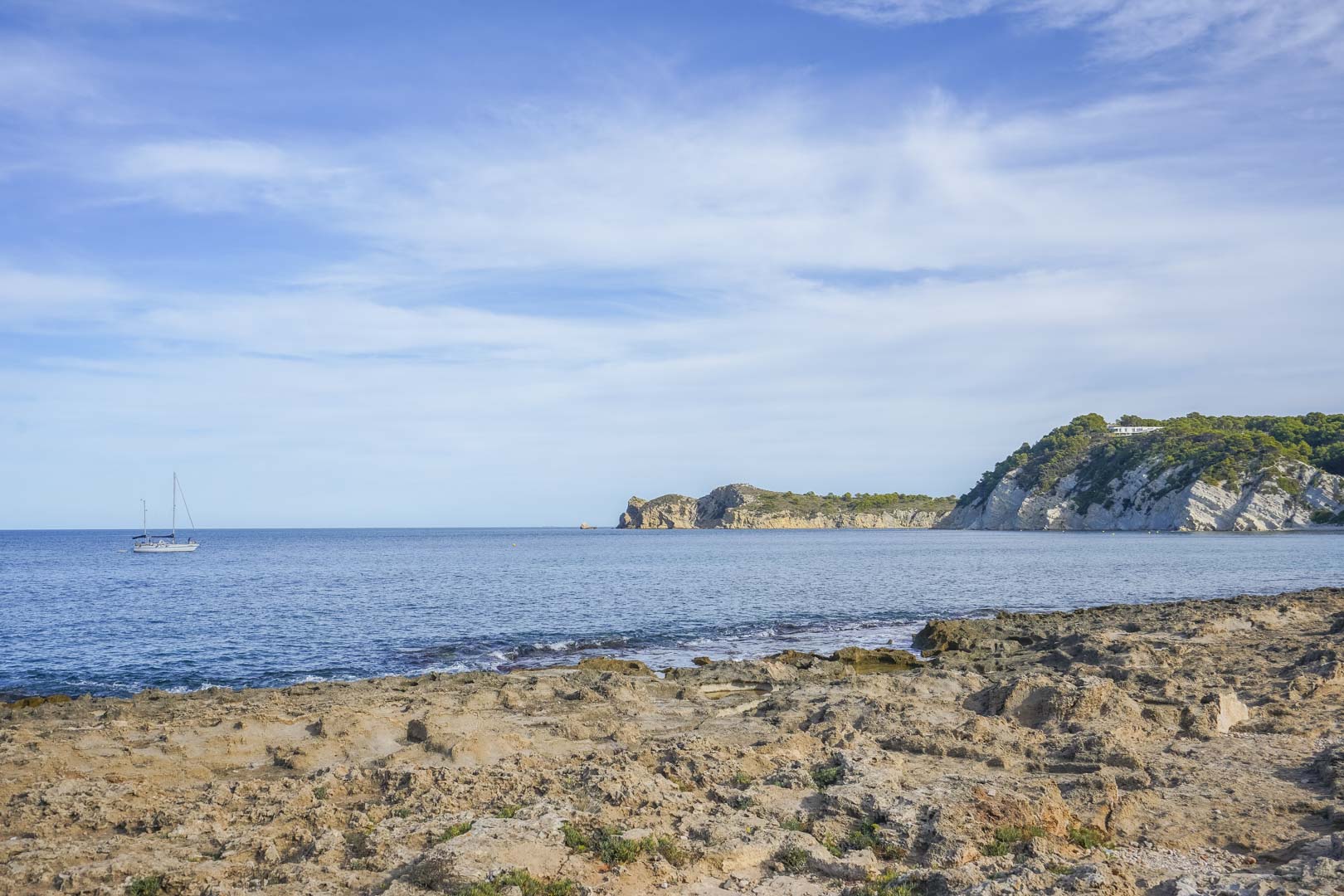 Vista de la costa rocosa de Jávea con el mar Mediterráneo, acantilados de piedra caliza y un velero navegando bajo un cielo despejado.