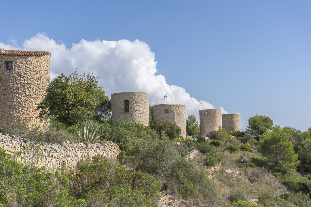 Vista de los históricos molinos de piedra de Jávea, situados en una colina con vegetación mediterránea autóctona y muros de piedra seca bajo un cielo despejado.