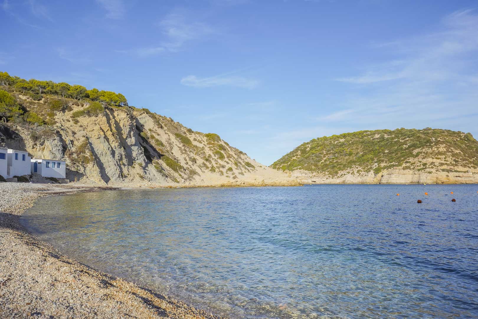 Vista de una cala de piedras en Jávea con aguas cristalinas, flanqueada por acantilados rocosos y vegetación mediterránea bajo un cielo despejado.