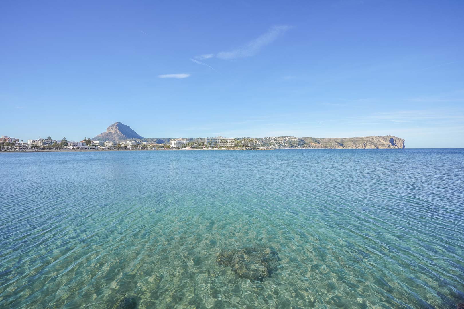 Vista panorámica de la bahía de Jávea con aguas cristalinas, destacando el monte Montgó al fondo y la costa del Arenal bajo un cielo despejado.