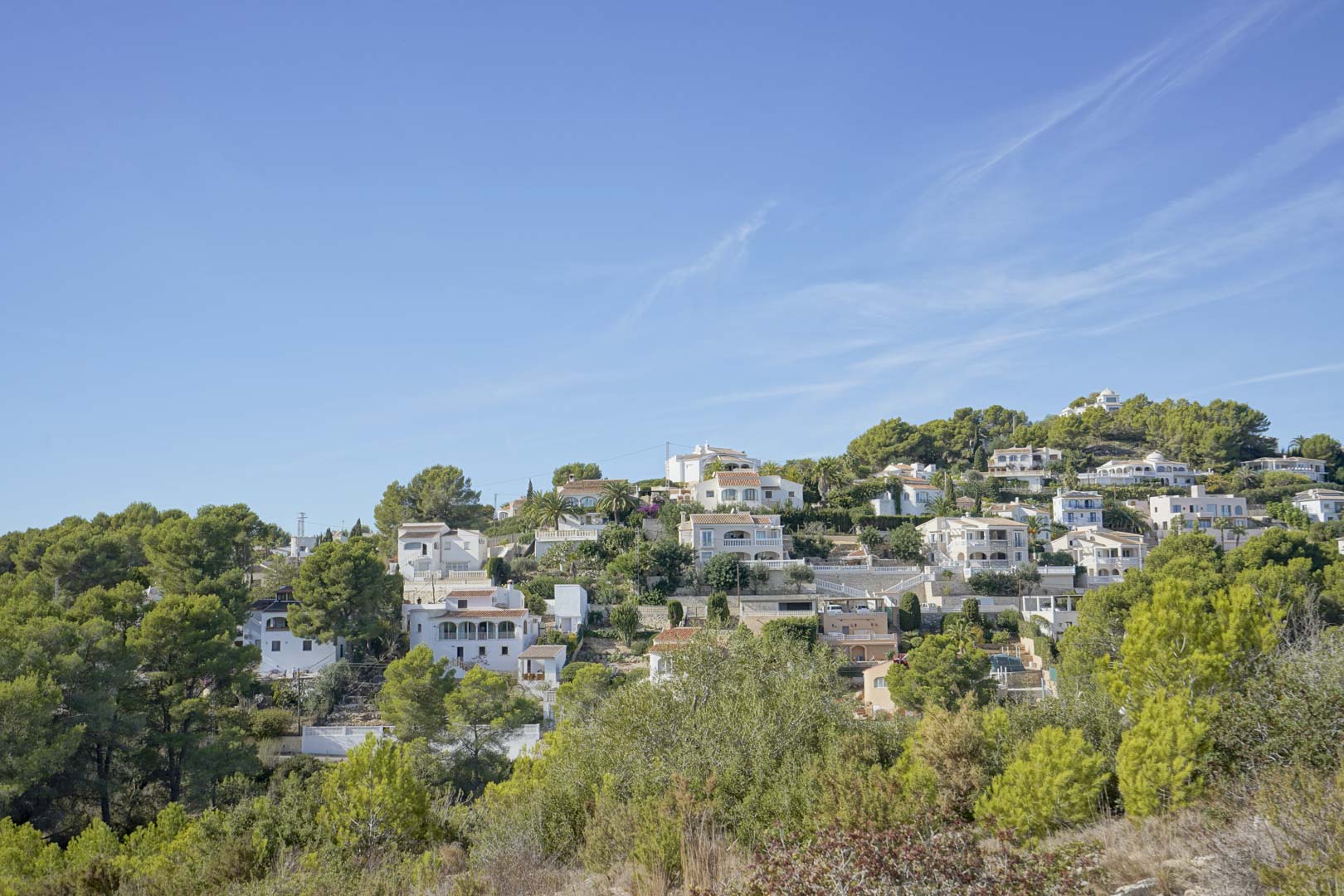 Vista panorámica de la urbanización Pinomar en Jávea, mostrando villas tradicionales integradas en la ladera arbolada bajo un cielo despejado.