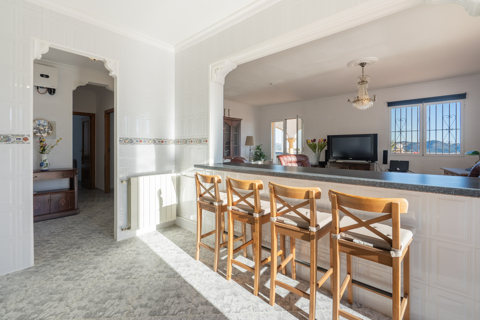 Cocina con barra americana y taburetes de madera abierta al salón, con paredes alicatadas, molduras clásicas y vistas a las montañas a través del ventanal.