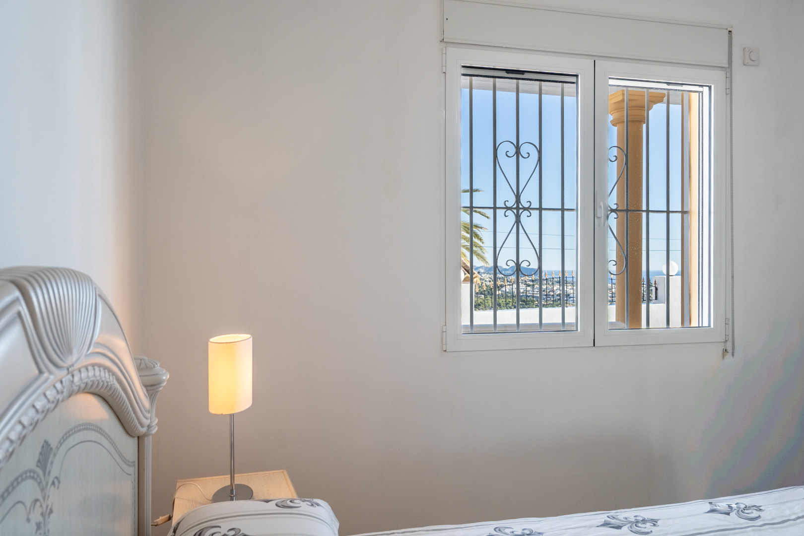 Dormitorio con cabecero tallado y ventana con rejas ornamentales que ofrece vistas despejadas hacia la costa de Calpe y el mar Mediterráneo.