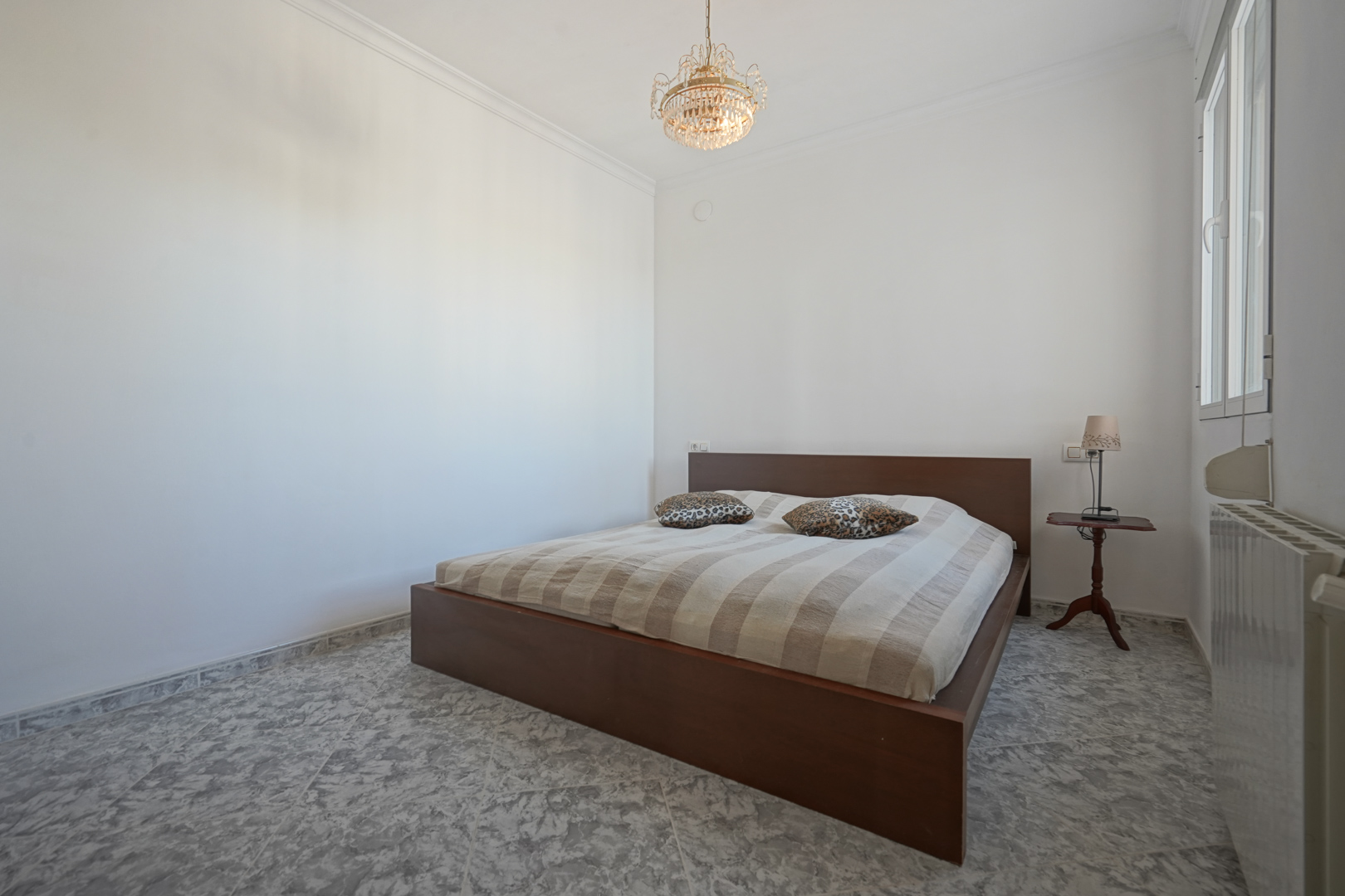 Dormitorio con cama de madera oscura, suelos de gres marmoleado, lámpara de araña clásica y radiador bajo la ventana lateral.