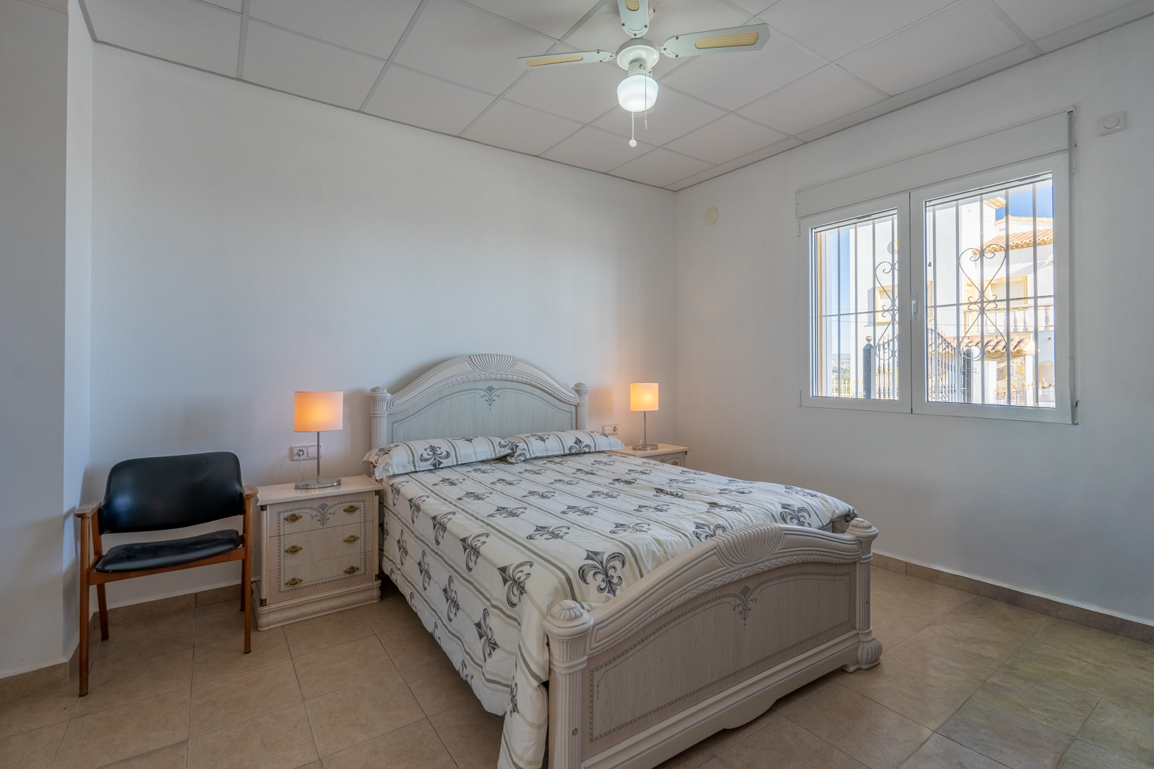Dormitorio con cama doble de estilo tradicional, suelos de gres, ventilador de techo y ventana con rejas de seguridad que aporta luz natural.