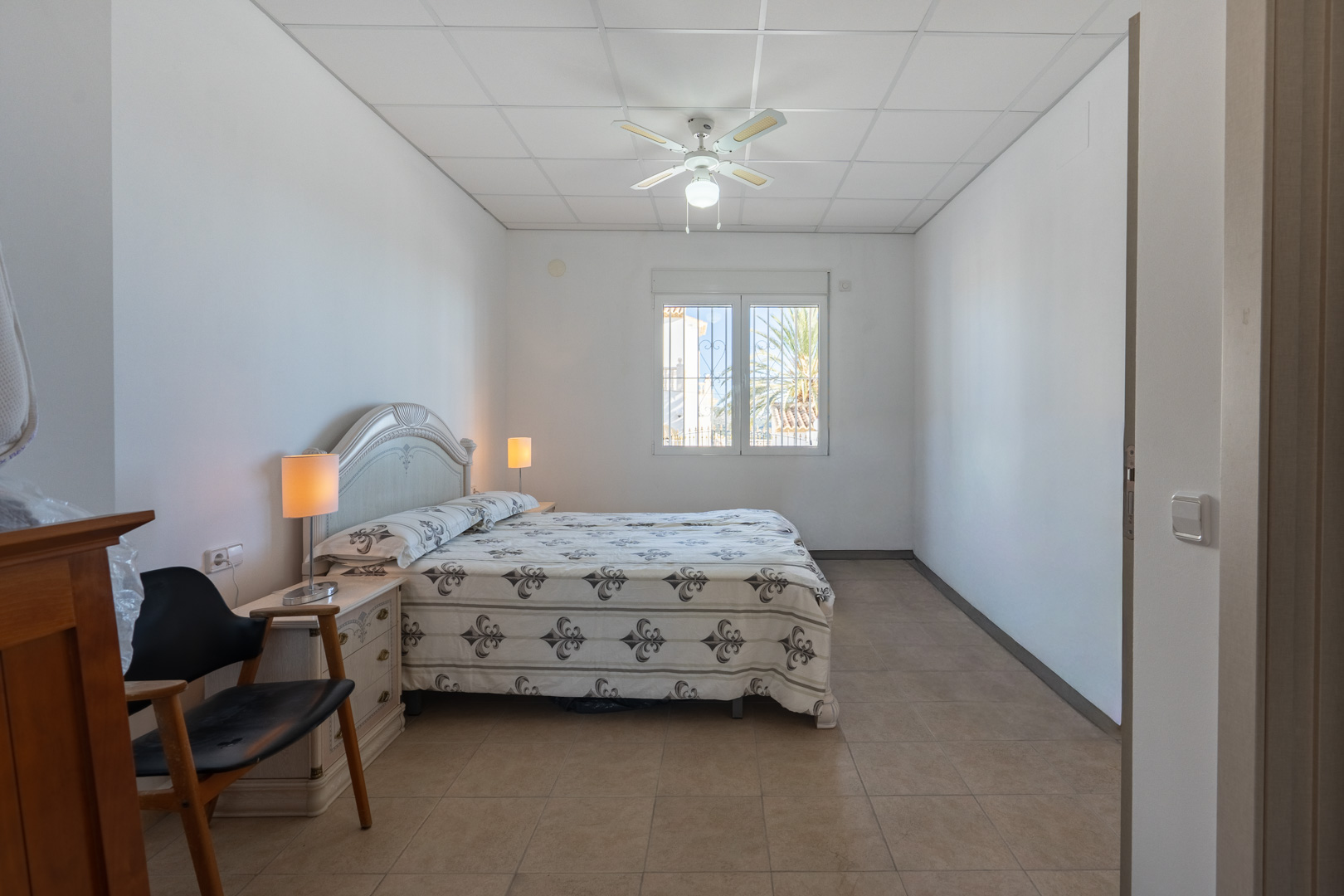 Dormitorio luminoso con cama doble, ventilador de techo y suelos cerámicos. Incluye una ventana con vistas al exterior y mobiliario de estilo clásico.