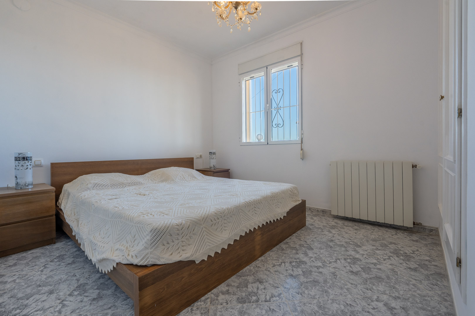 Dormitorio luminoso con suelos cerámicos de efecto mármol, cama de madera con mesitas a juego, armarios empotrados blancos y calefacción central por radiador.