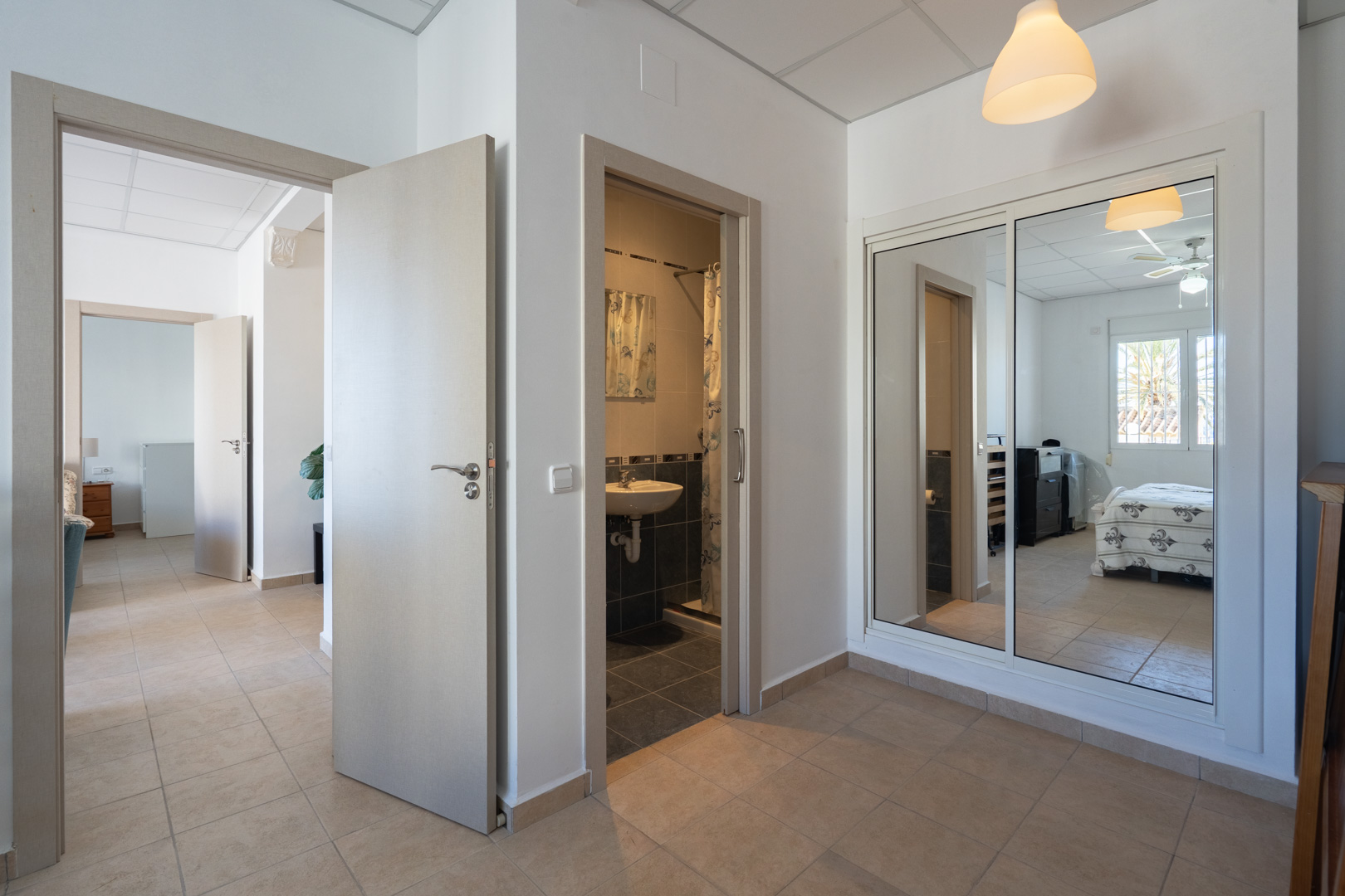 Pasillo de distribución con suelos cerámicos, armario empotrado de puertas espejadas y acceso directo a un cuarto de baño y un dormitorio con ventilador de techo.