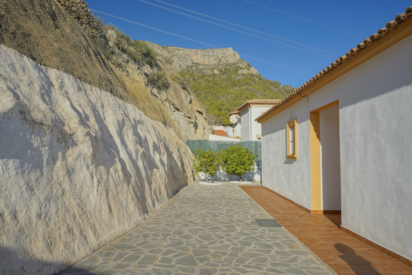 Pasillo exterior con pavimento de piedra irregular junto a una pared de roca natural, con la Sierra de Oltà al fondo y árboles cítricos.