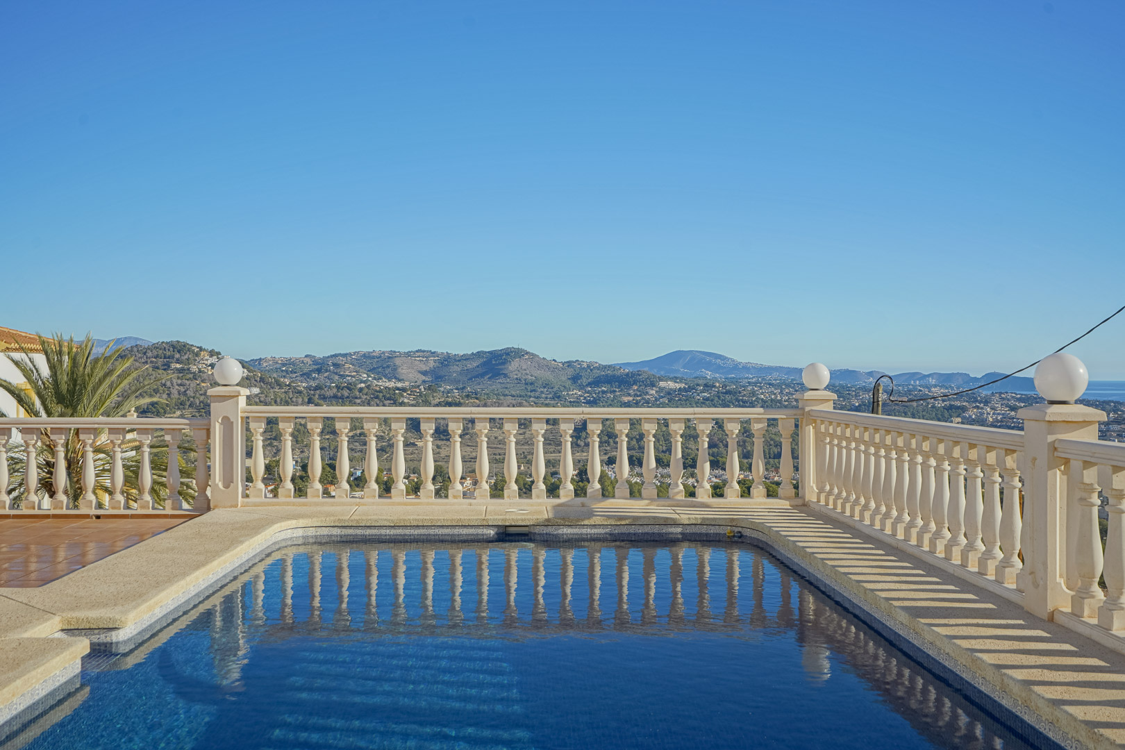 Piscina rectangular con balaustrada de piedra clásica y vistas panorámicas hacia la costa de Moraira y las montañas circundantes bajo un cielo despejado.