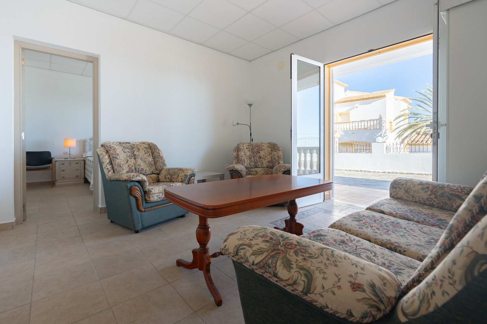 Salón del apartamento de invitados con mobiliario floral, mesa de madera y acceso directo a la terraza. Se aprecia el dormitorio adyacente y suelos cerámicos.