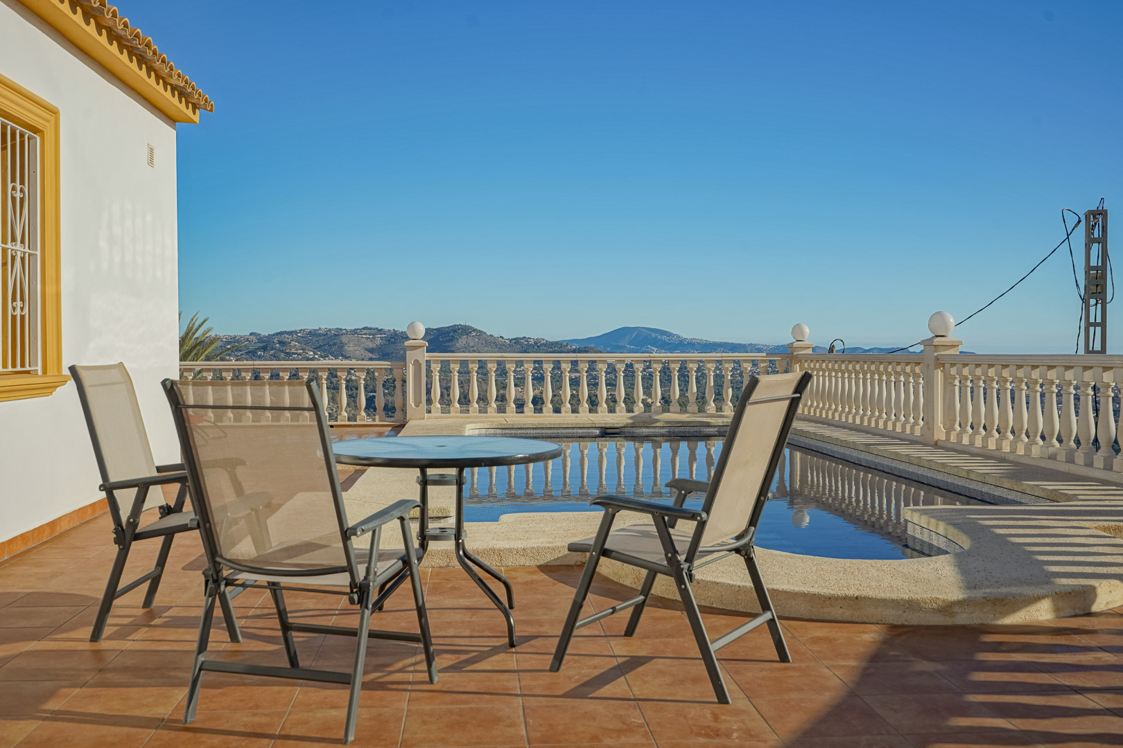 Terraza exterior con baldosas de terracota, zona de estar con mesa de cristal y vistas panorámicas a las montañas y la piscina privada con balaustrada clásica.