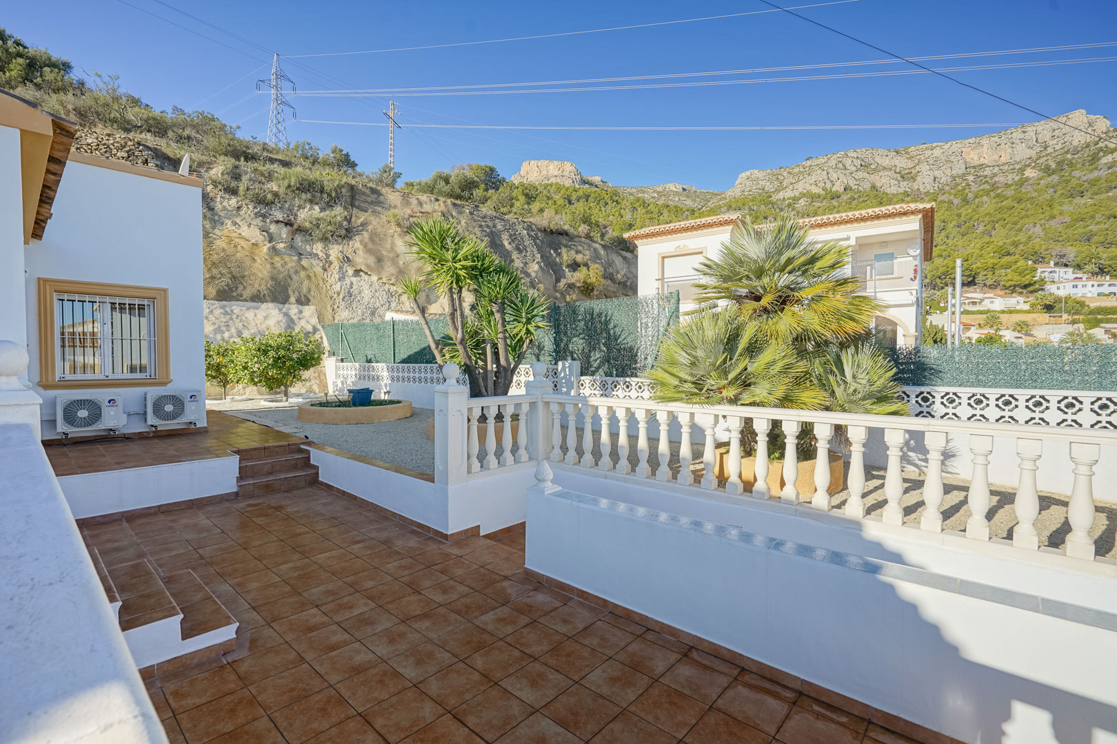 Terraza exterior con suelos de gres rústico y balaustrada blanca, rodeada de un jardín de bajo mantenimiento con palmeras y vistas a la Sierra de Oltà.