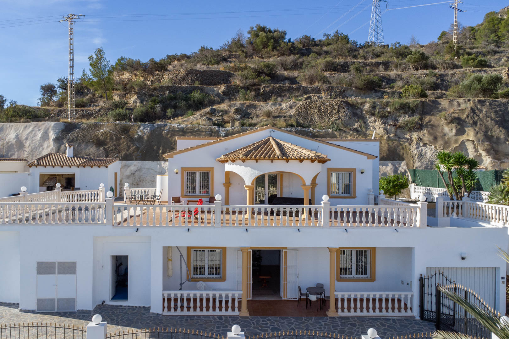 Vista exterior de la villa de dos plantas con fachada blanca, balaustradas clásicas y porches cubiertos, situada frente a la ladera de la Sierra de Oltà.