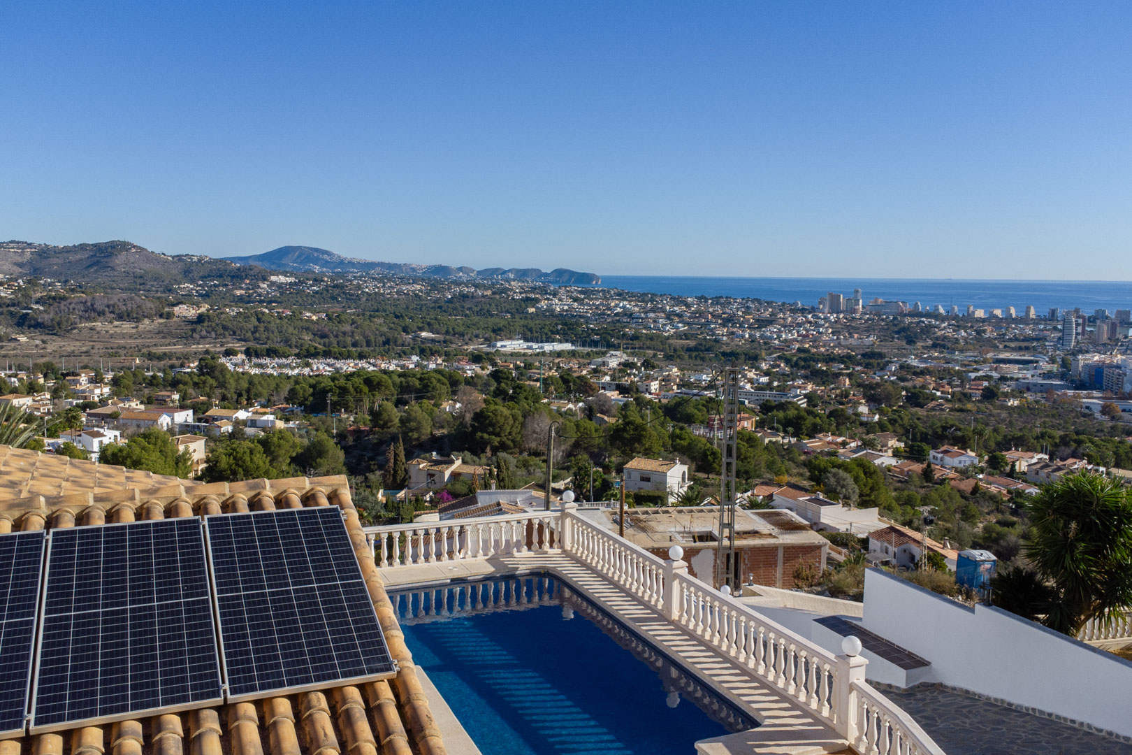 Vista panorámica desde la villa que muestra la piscina con balaustrada blanca, paneles solares sobre tejado de teja y el skyline de Calpe frente al mar Mediterráneo.