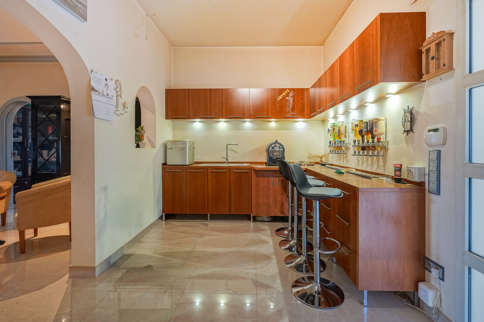 Bar integrado con armarios de madera, fregadero y dispensadores de pared. Dispone de taburetes metálicos, iluminación LED bajo los muebles y suelos cerámicos pulidos.
