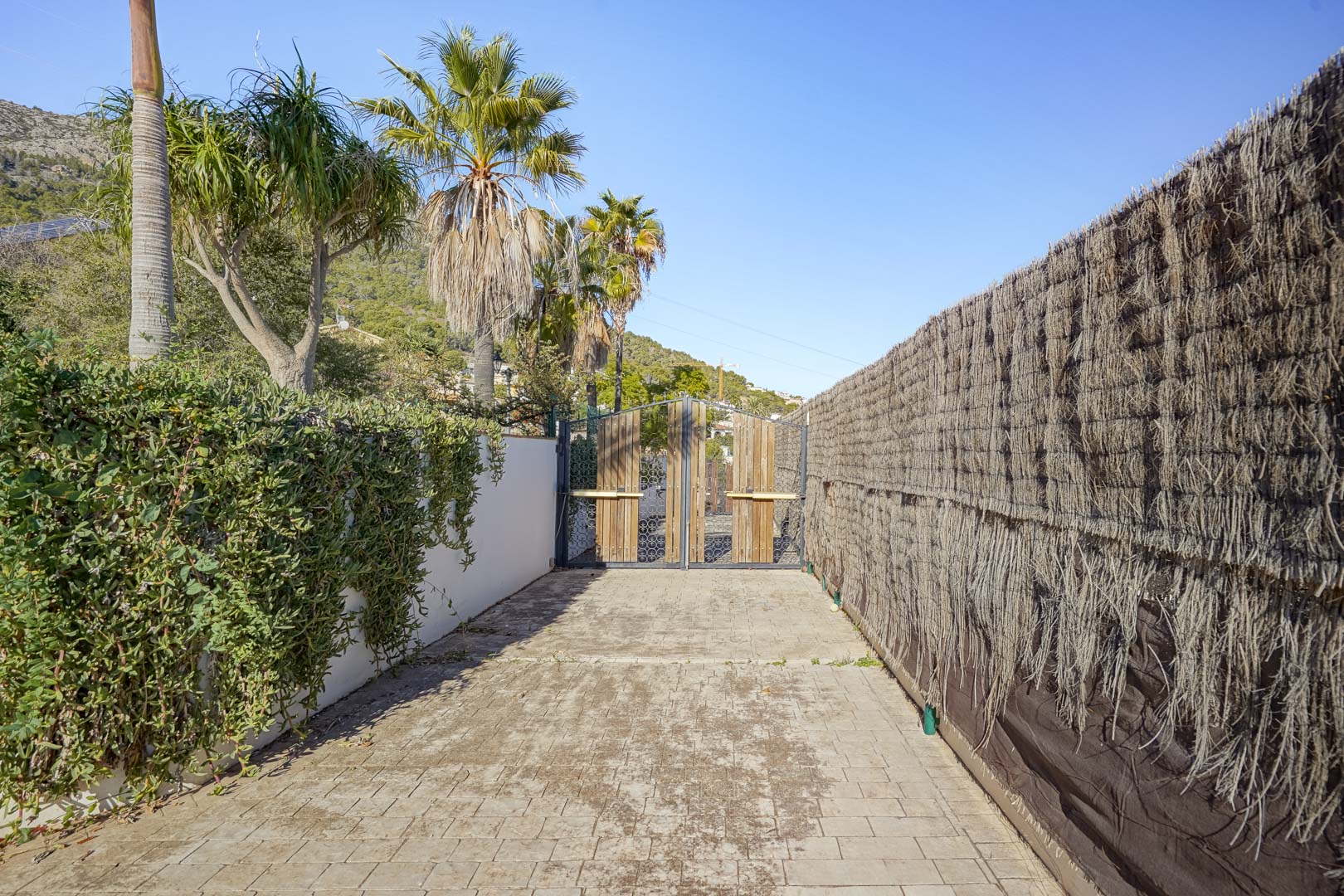 Entrada a la propiedad con camino pavimentado y puerta de madera y forja, flanqueada por vegetación mediterránea, palmeras y muros con cerramiento de brezo.