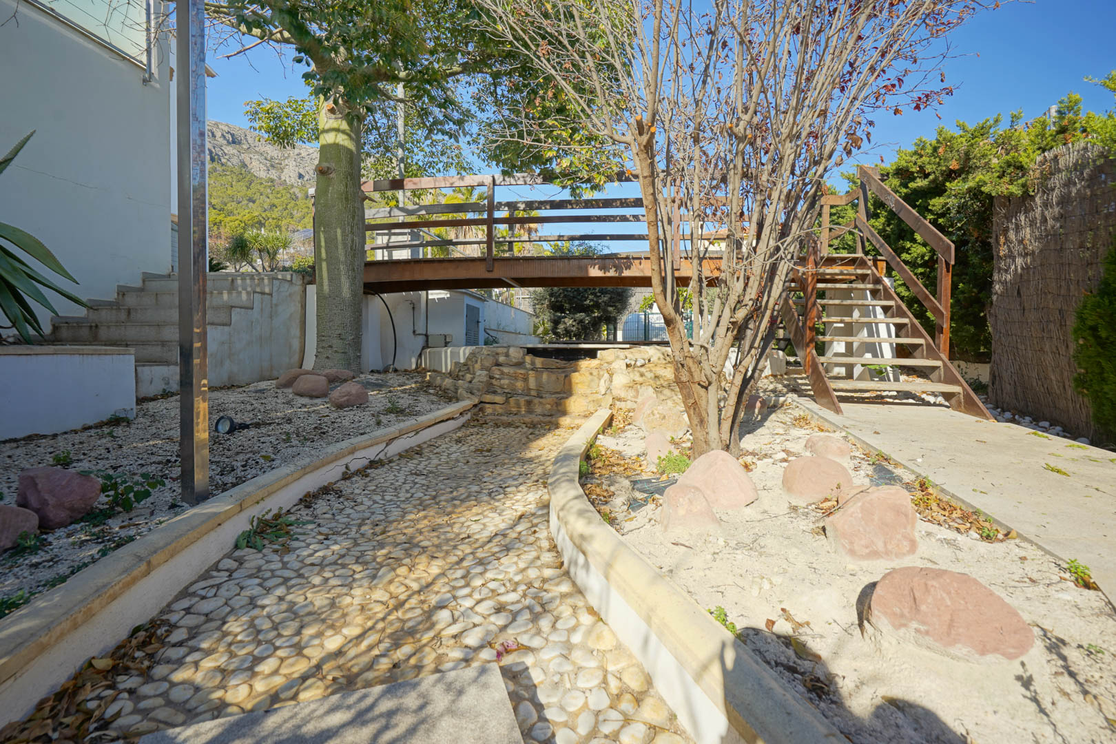 Jardín con pasarela de madera sobre cascada decorativa de piedra, camino empedrado y escaleras de acceso, con vistas a la Sierra de Oltamar al fondo.