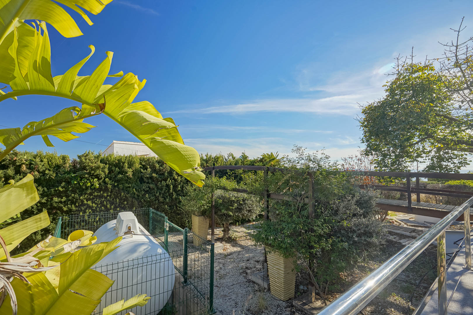 Jardín lateral con depósito de gas vallado, pasarela de madera y vegetación tropical que incluye grandes hojas de banano.