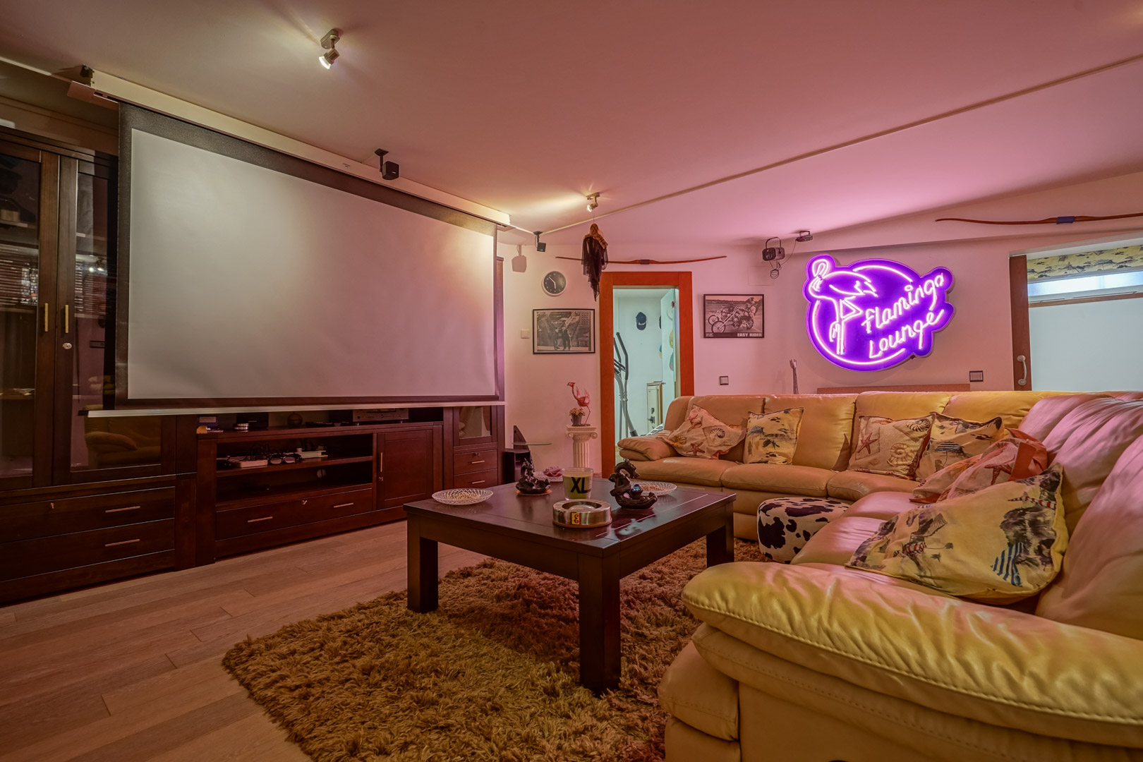 Sala multimedia equipada con pantalla de proyección retráctil, mueble de madera oscura a medida y sofá rinconero de cuero. Destaca su iluminación ambiental con letrero de neón.