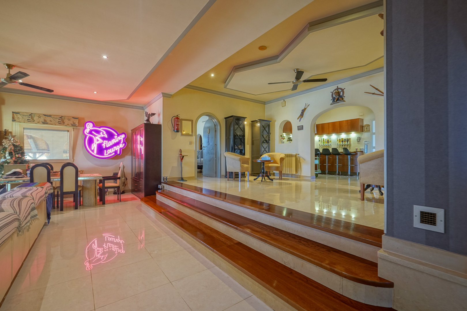 Salón de concepto abierto con niveles diferenciados por escalones de madera, suelos de mármol pulido y una zona de bar integrada bajo arcos decorativos.