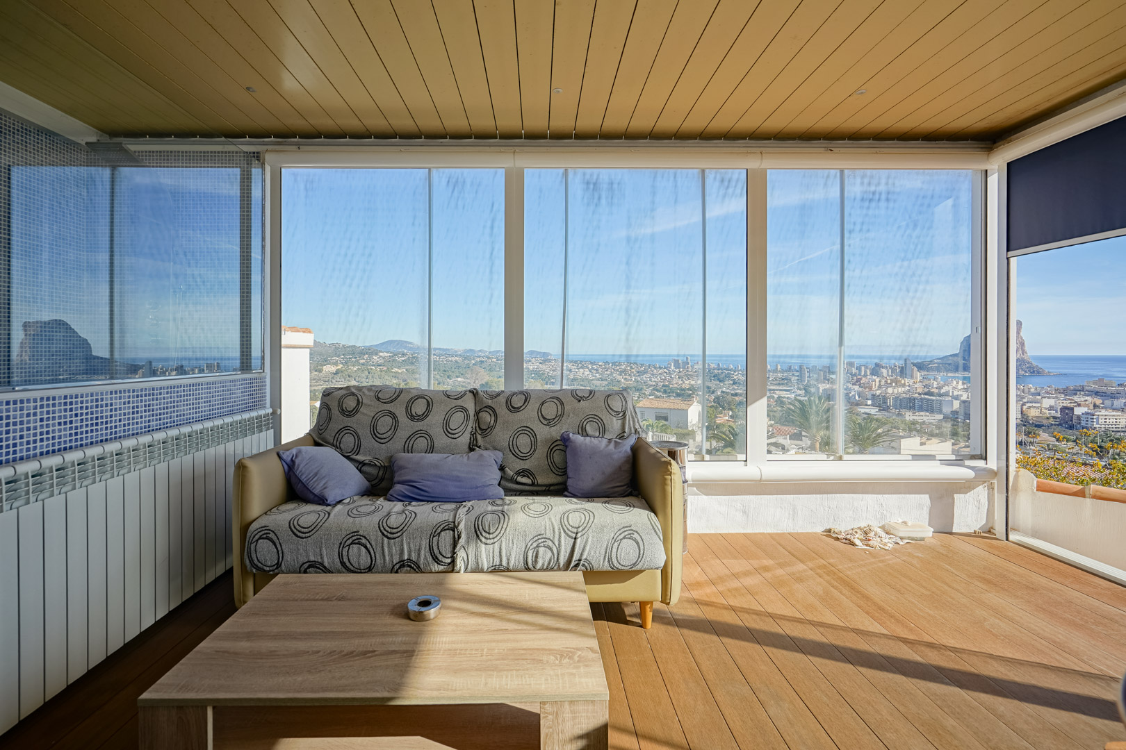 Terraza acristalada con cerramiento de vidrio, suelos de madera y vistas panorámicas al skyline de Calpe y el Peñón de Ifach. Incluye techo panelado y zona de estar.