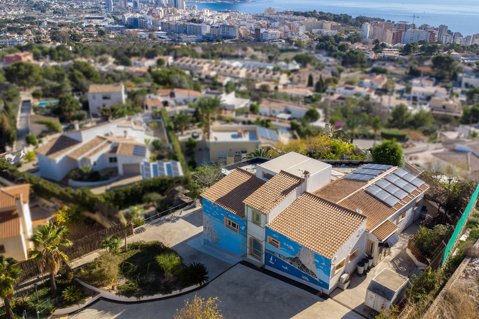 Vista aérea de la villa con murales artísticos, tejado de teja cerámica y sistema de paneles solares, ubicada en una zona residencial con vistas al mar y al skyline de Calpe.