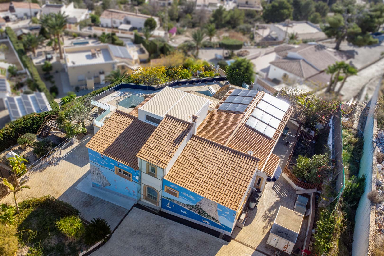 Vista aérea de la villa que muestra el tejado de teja tradicional, la instalación de paneles solares y un mural artístico detallado en la fachada lateral.