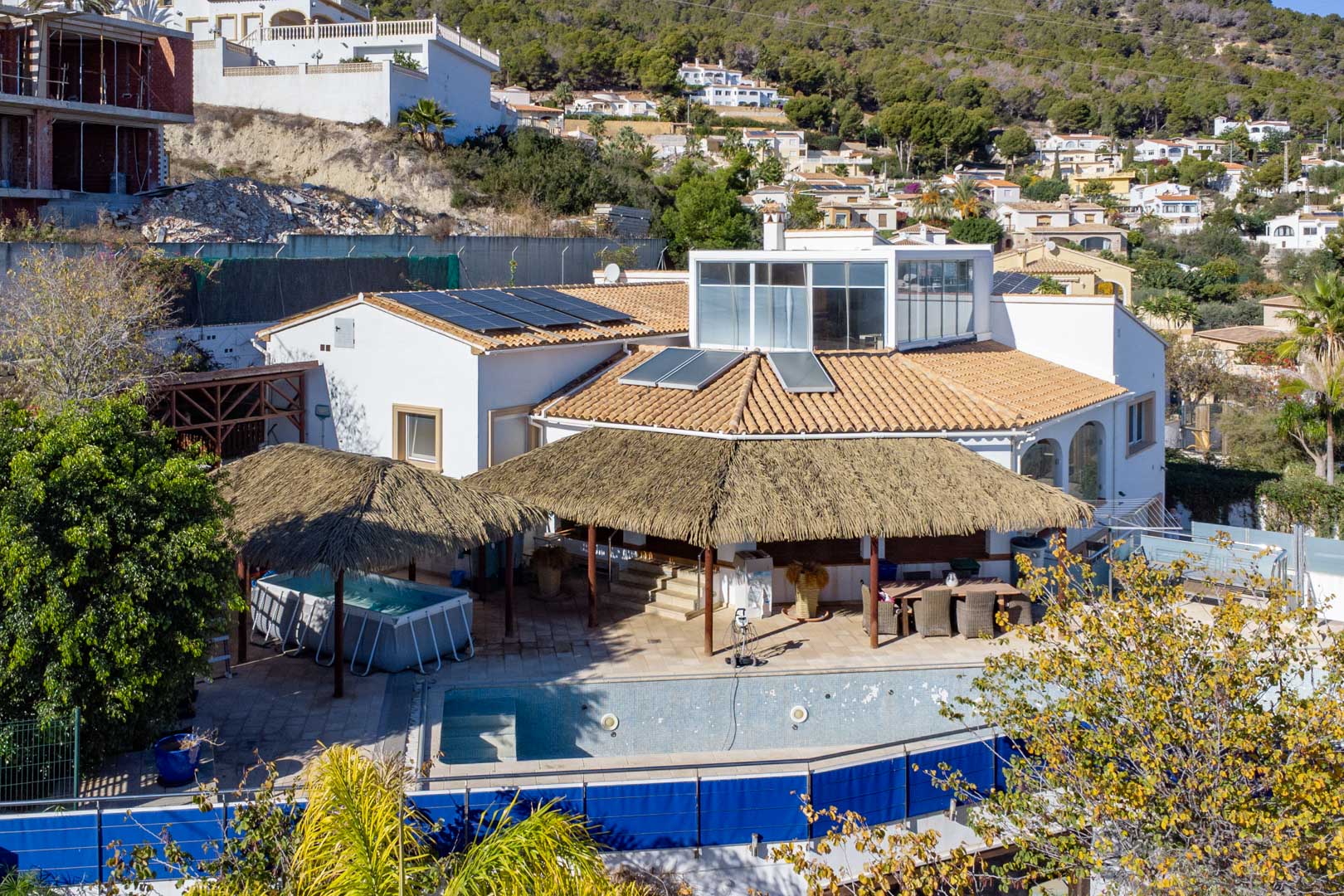 Vista exterior de la villa con paneles solares, terrazas con pérgolas de brezo y zona de piscina, destacando el cerramiento acristalado superior en la ladera de Calpe.