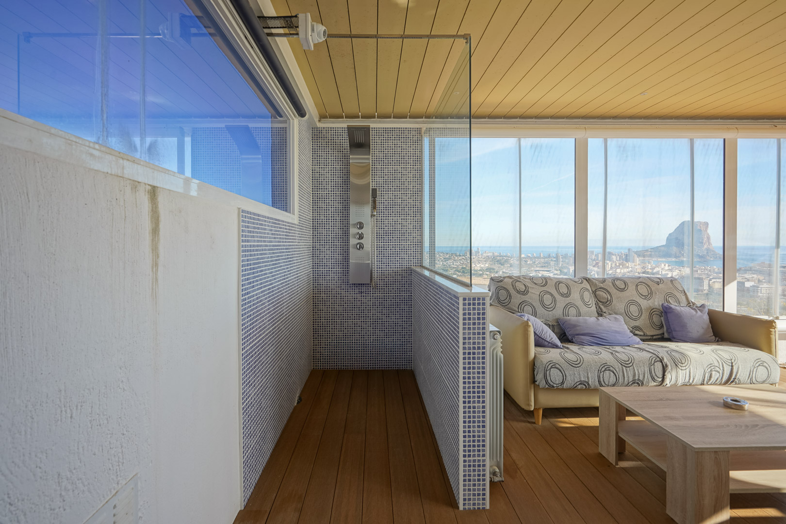 Zona de bienestar en planta superior con ducha de hidromasaje, gresite azul y cerramiento acristalado con vistas panorámicas al Peñón de Ifach y la costa de Calpe.