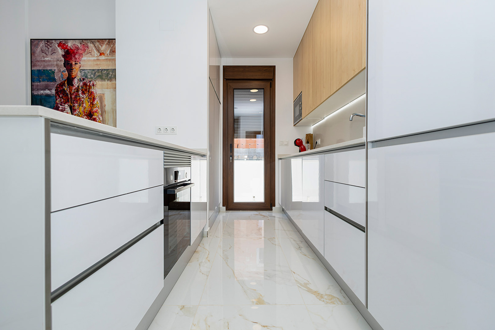 Cocina moderna con armarios blancos de alto brillo, muebles superiores en madera y suelo porcelánico efecto mármol. Incluye electrodomésticos integrados y acceso directo al exterior.
