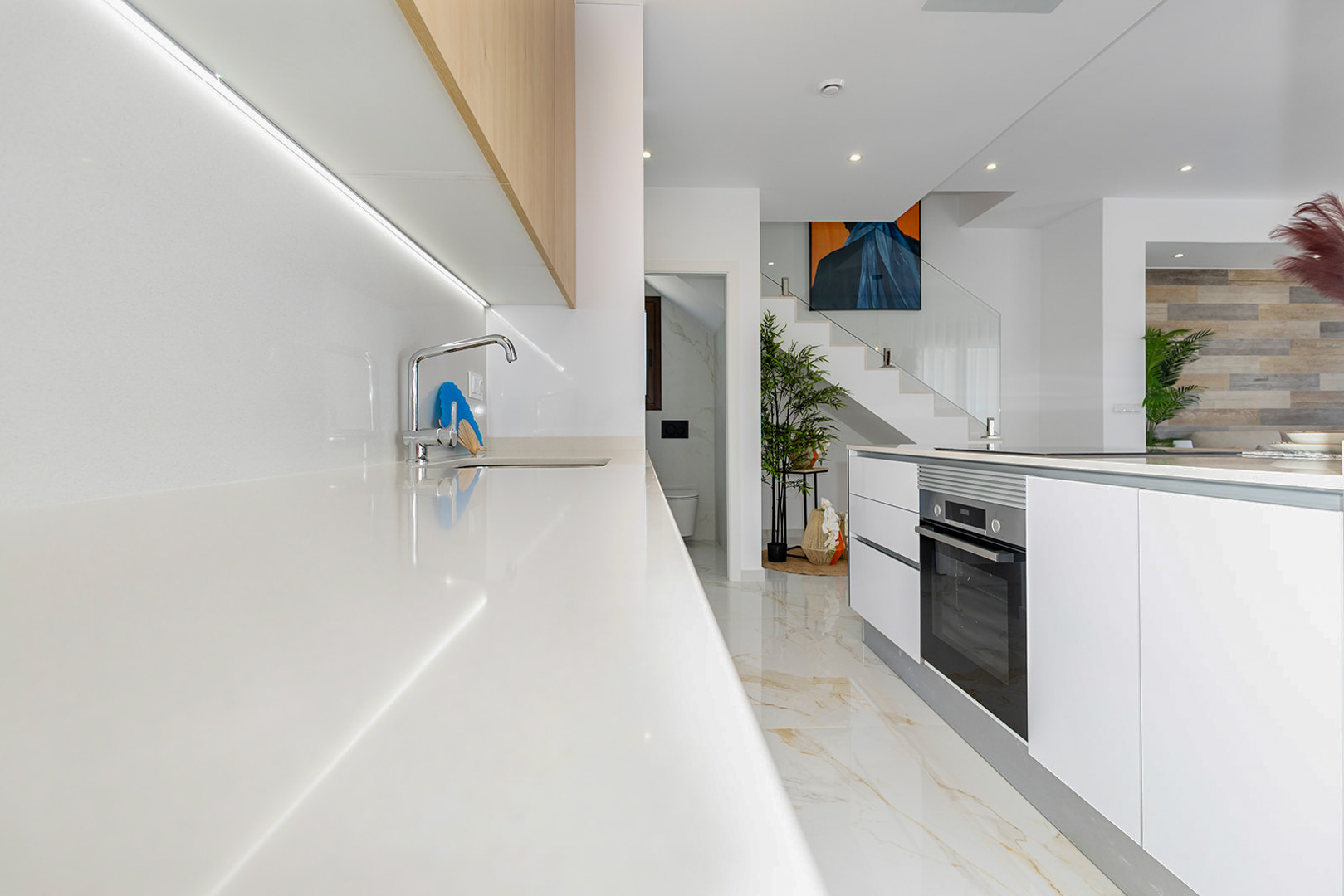Cocina moderna con encimera blanca, iluminación LED bajo muebles de madera y una isla con horno integrado. Al fondo destaca la escalera con barandilla de cristal.
