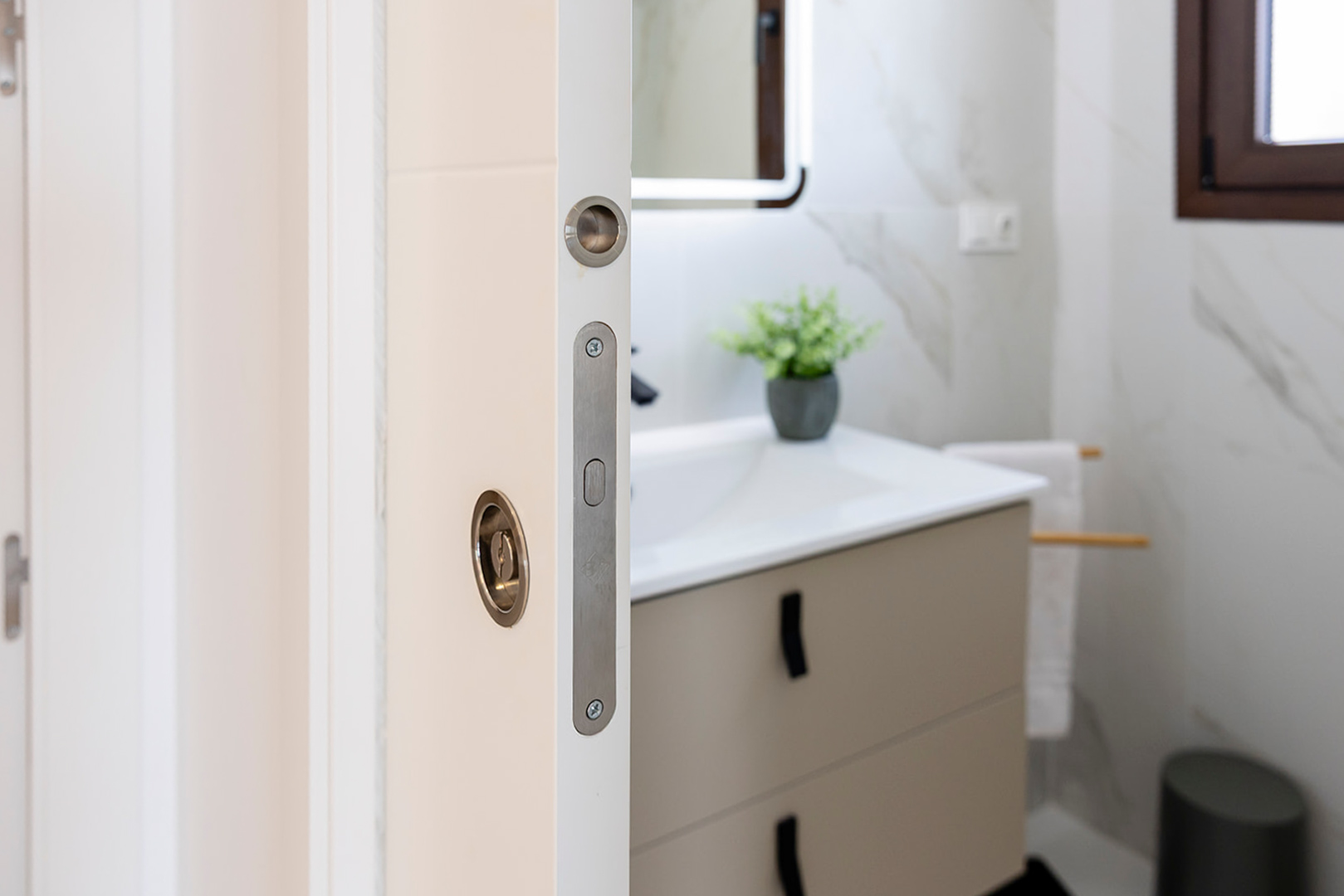 Detalle de puerta corredera blanca con herrajes metálicos integrados, que da acceso a un baño moderno con revestimiento cerámico marmoleado y mueble de lavabo suspendido.
