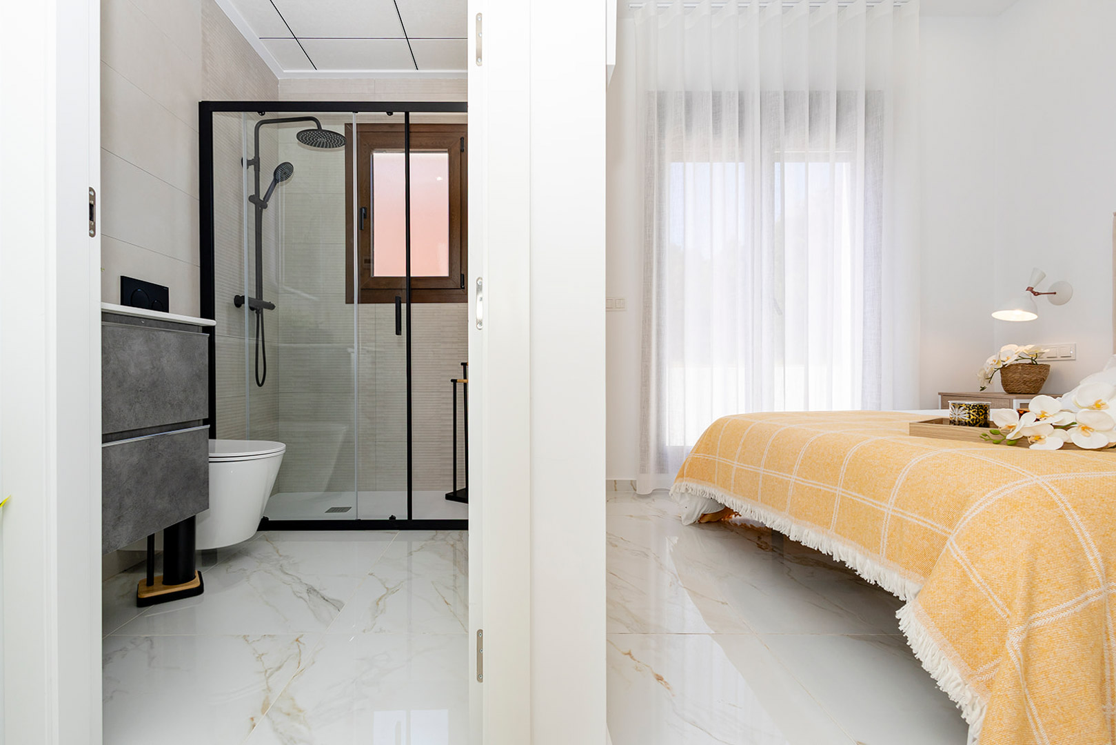 Dormitorio con baño en suite integrado, suelos de gres porcelánico efecto mármol y ducha con perfilería negra y mampara de cristal.