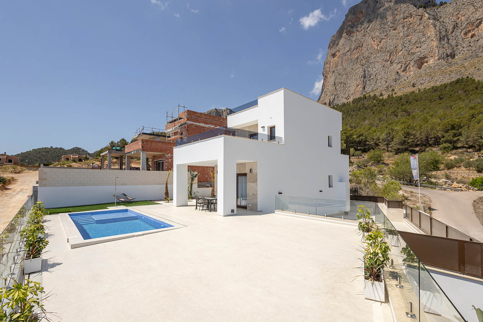 Exterior de villa moderna con fachada blanca, piscina rectangular de gresite azul y amplia terraza de gres porcelánico bajo la falda de una montaña rocosa.