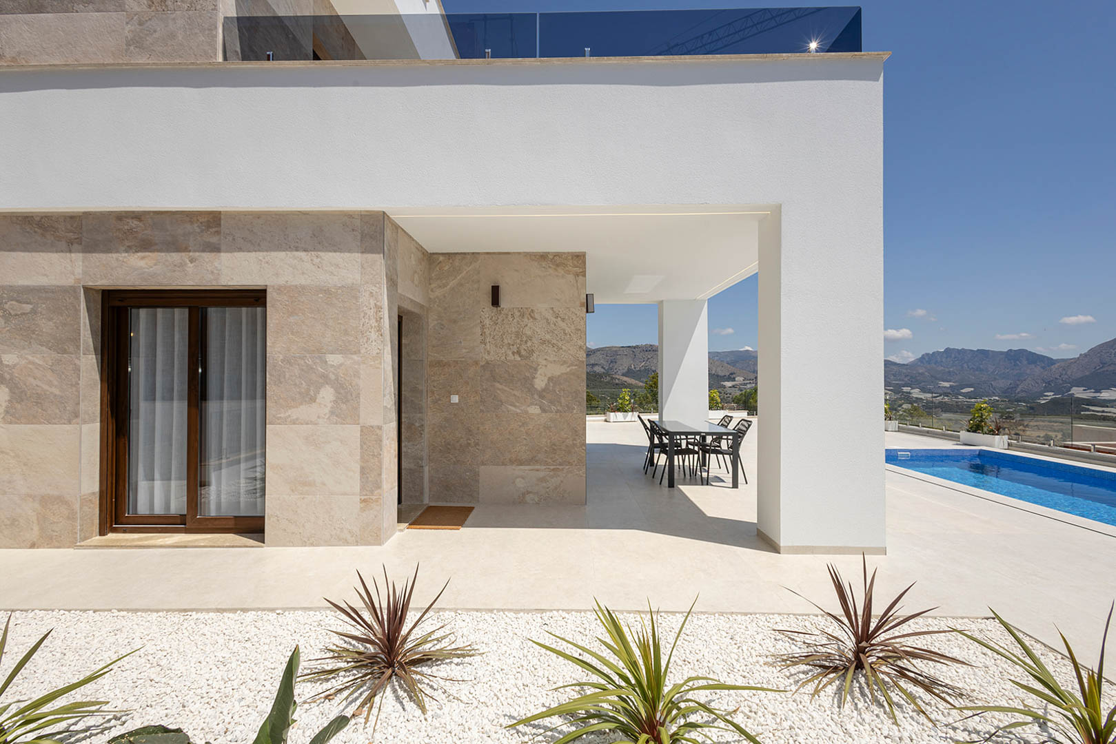 Fachada de diseño moderno con revestimiento de piedra natural, porche cubierto con zona de comedor, piscina privada y vistas despejadas a las montañas.