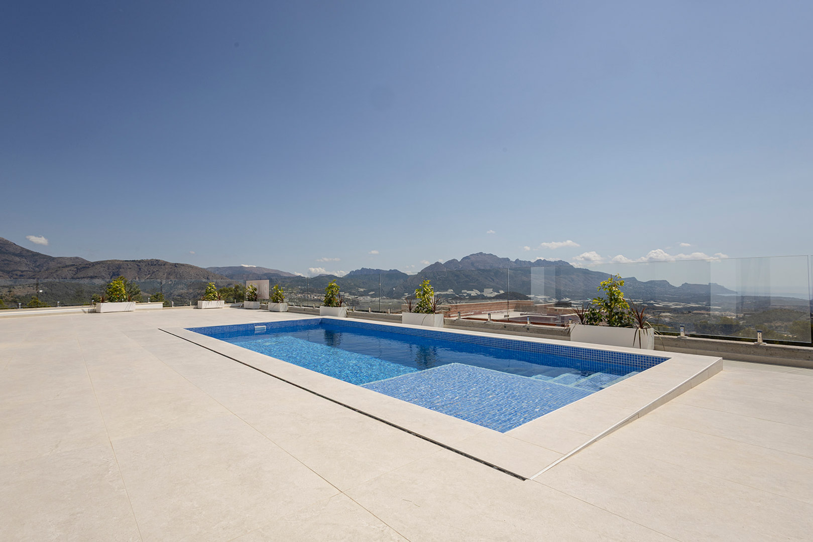 Piscina privada de gresite azul integrada en una amplia terraza de gres porcelánico claro, con barandilla de cristal y vistas panorámicas a la montaña y el mar.