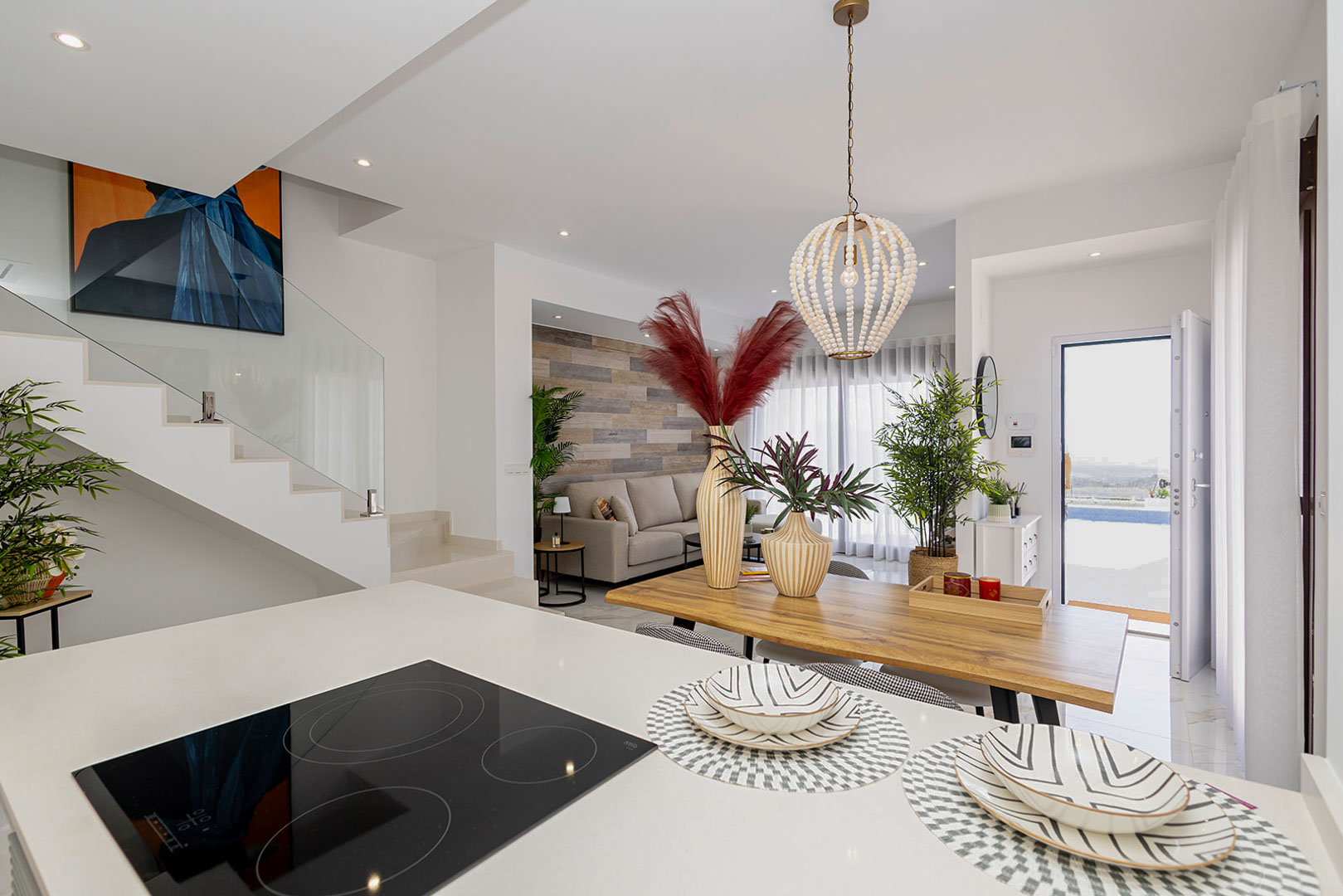 Salón-comedor integrado con cocina abierta, destacando la escalera con barandilla de cristal, placa de inducción en isla y acceso directo al exterior.
