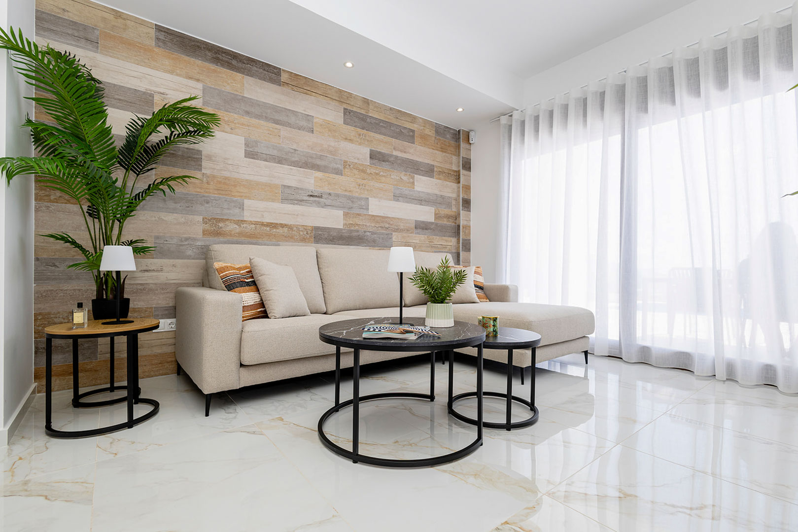 Salón con pared de acento en lamas de madera, suelo de gres porcelánico efecto mármol y sofá beige junto a grandes ventanales con cortinas traslúcidas.