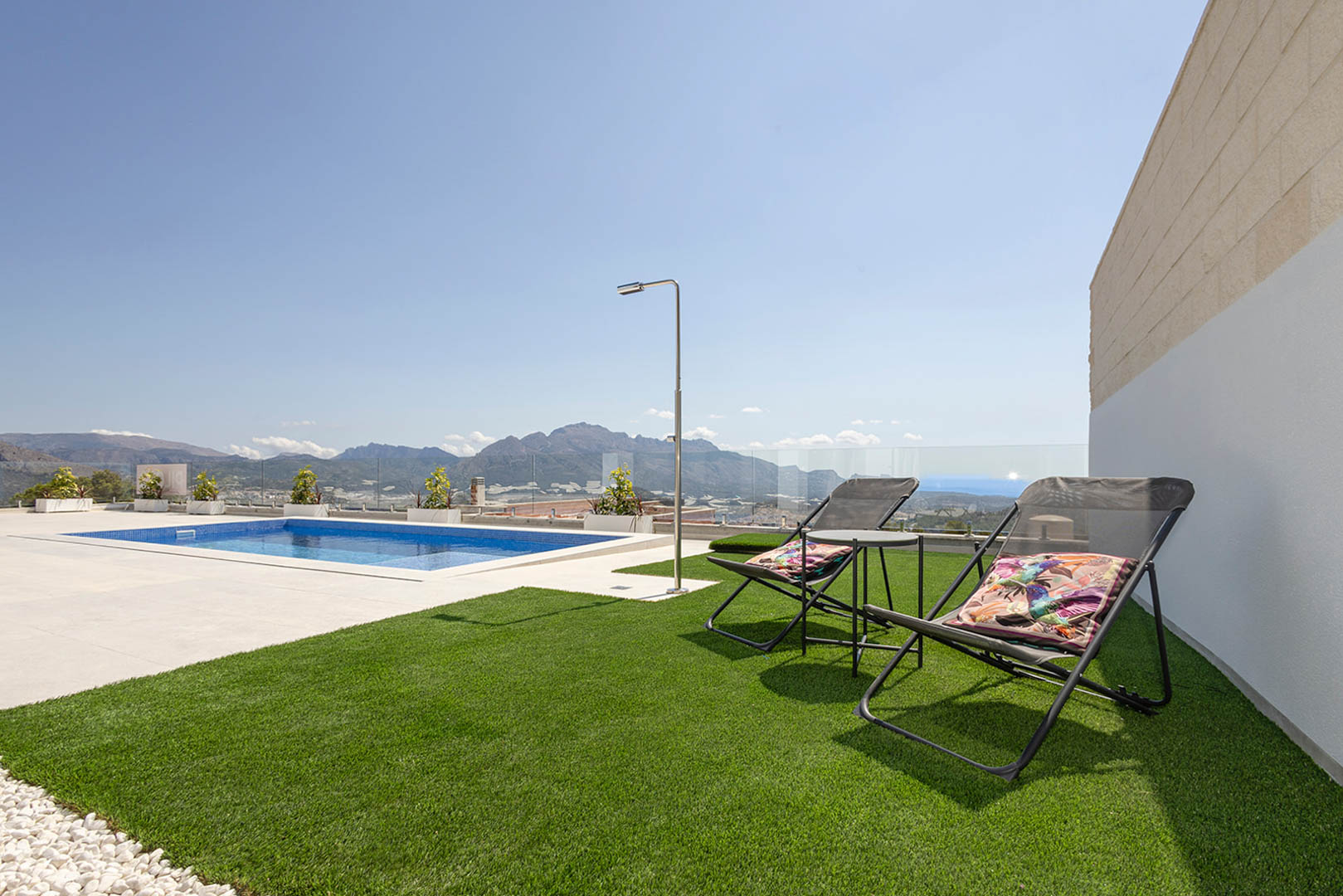 Terraza exterior con piscina de mosaico azul, zona de césped artificial con tumbonas y ducha de acero inoxidable. Vistas panorámicas a la montaña protegidas por barandillas de cristal.