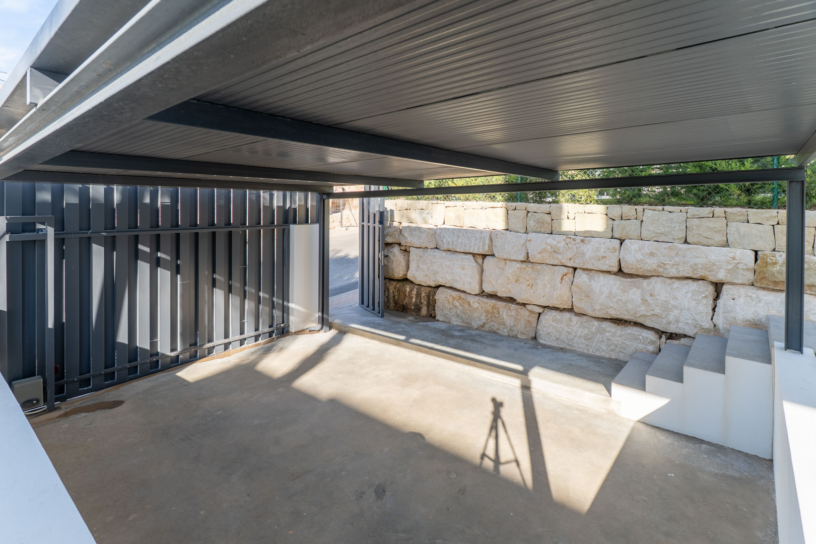 Aparcamiento privado cubierto con estructura metálica y suelo de hormigón, delimitado por un muro de piedra natural y acceso mediante portón automatizado.