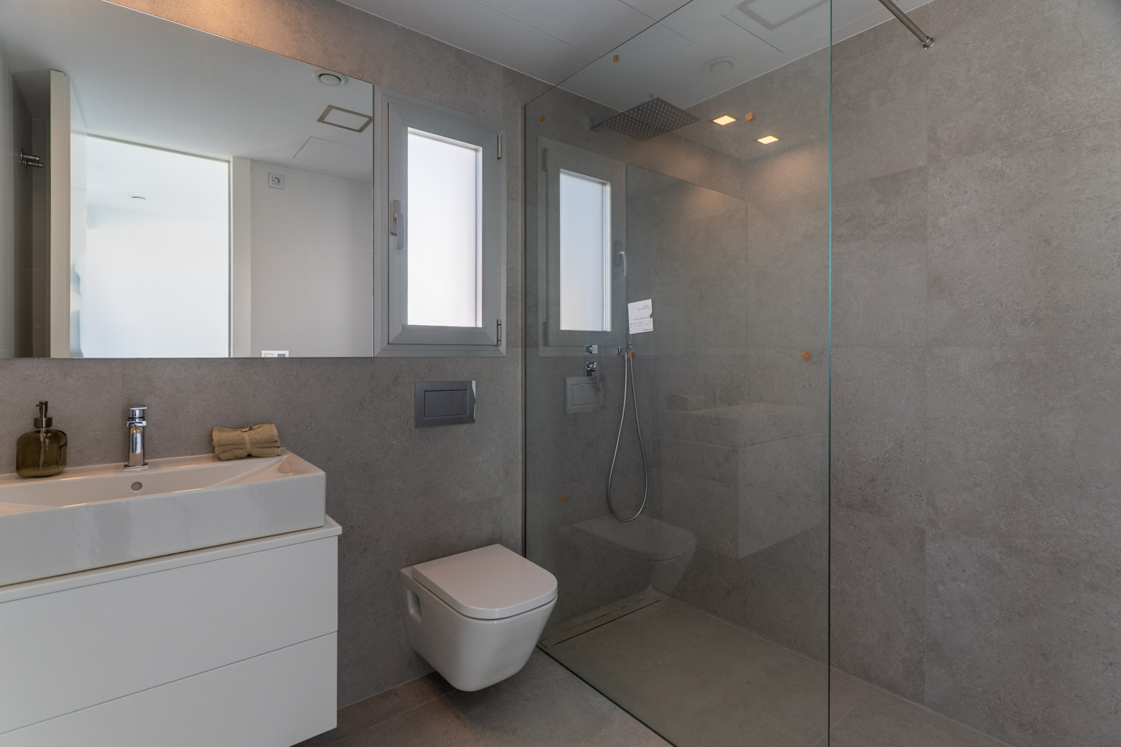Baño contemporáneo con azulejos grises, ducha de obra con mampara de vidrio, sanitario suspendido y lavabo integrado sobre mueble blanco minimalista.