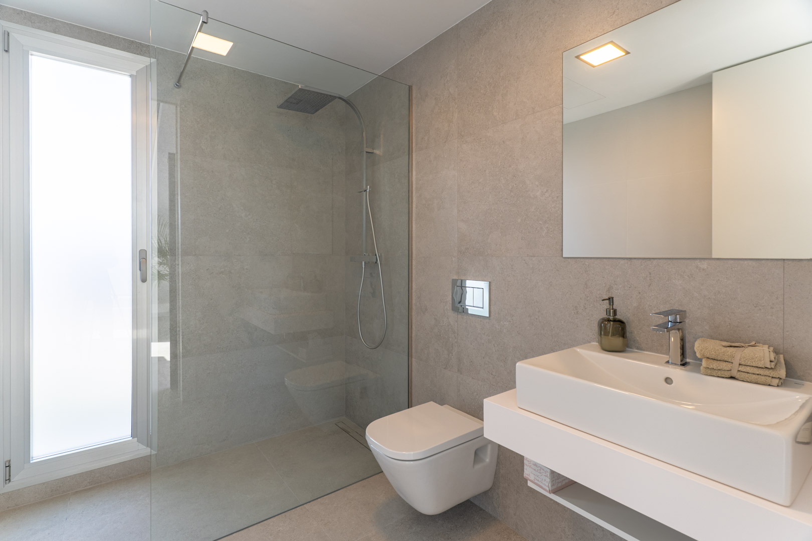 Baño moderno con revestimiento cerámico en tonos grises, ducha a ras de suelo con mampara de cristal, inodoro suspendido y lavabo de diseño sobre encimera blanca.