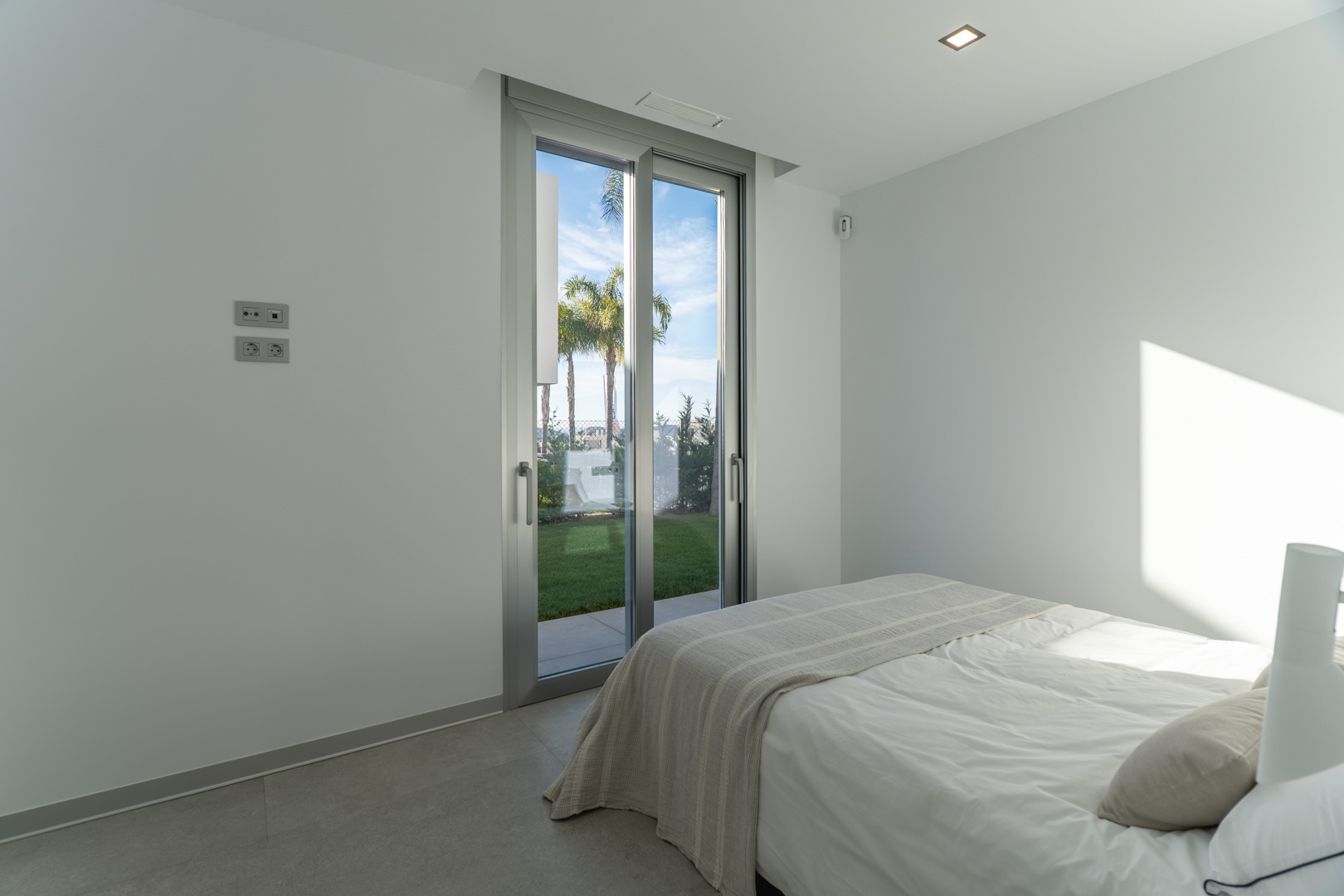 Dormitorio luminoso con acceso directo al jardín mediante puerta corredera de aluminio, suelos de gres porcelánico y paredes blancas minimalistas.