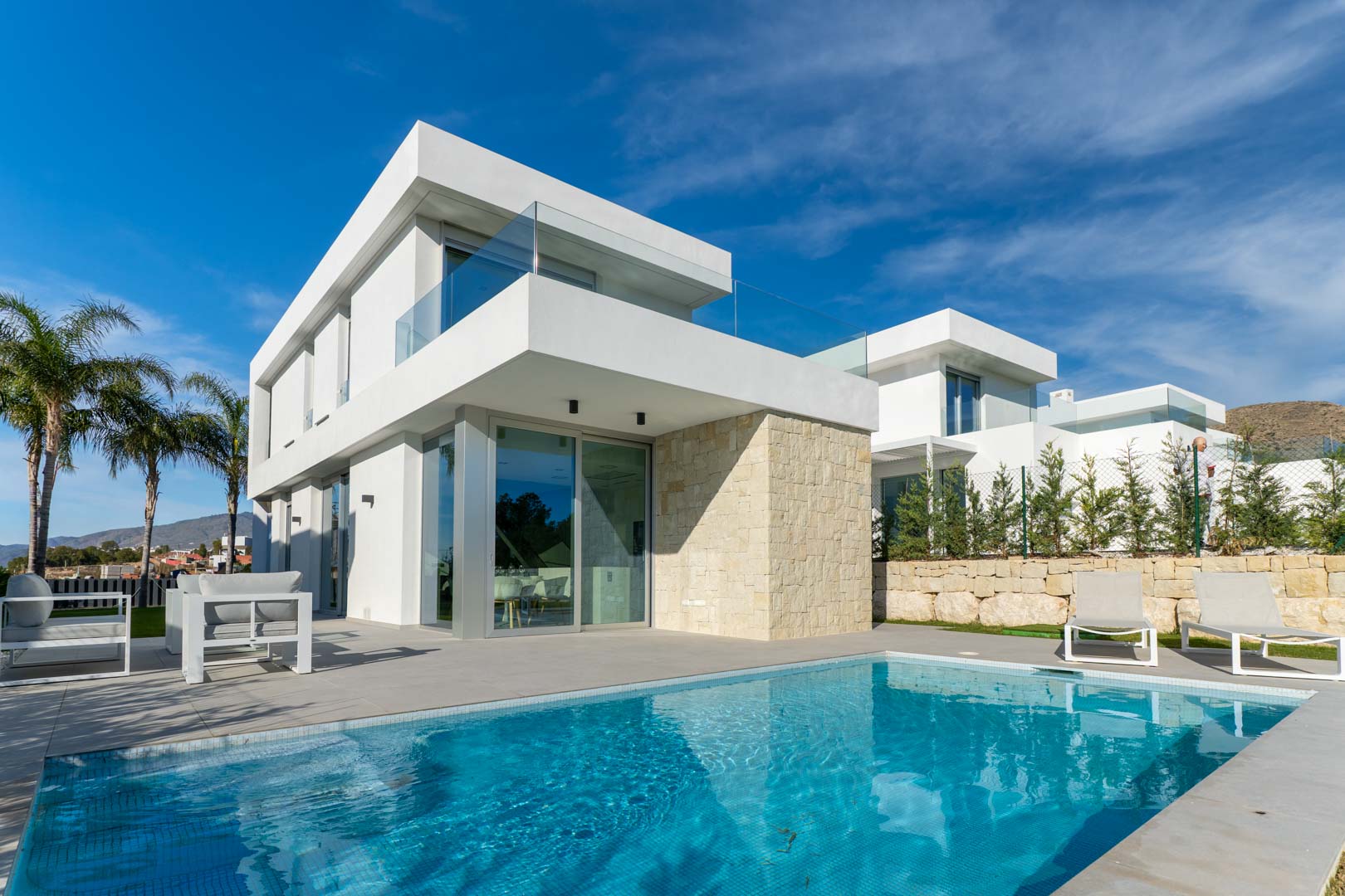 Exterior de villa moderna con piscina privada, fachada blanca minimalista, muro de piedra natural y terrazas con barandillas de cristal.