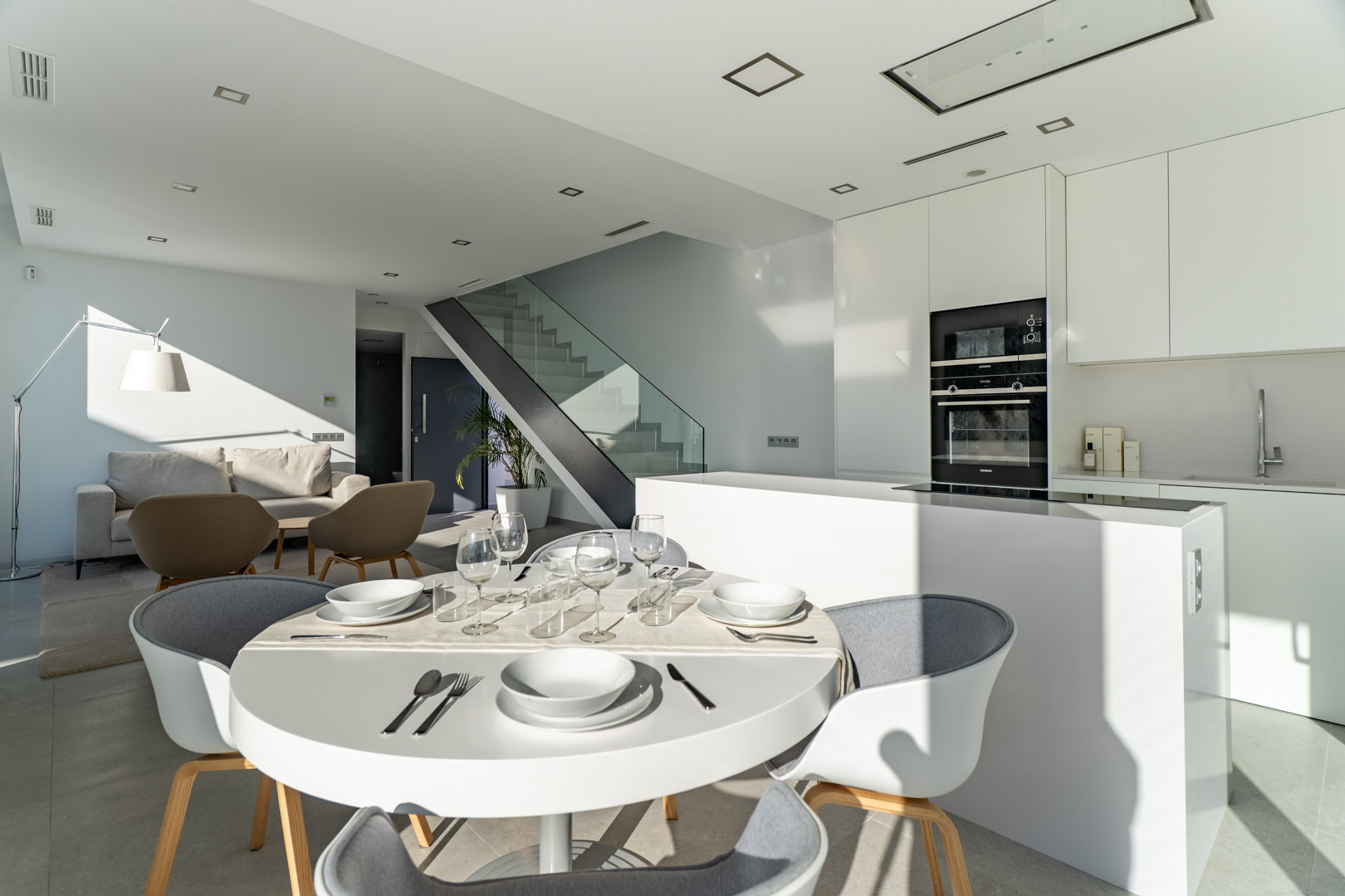 Salón-comedor de concepto abierto con cocina minimalista blanca, isla central y electrodomésticos integrados. Incluye una escalera de diseño con barandilla de cristal.